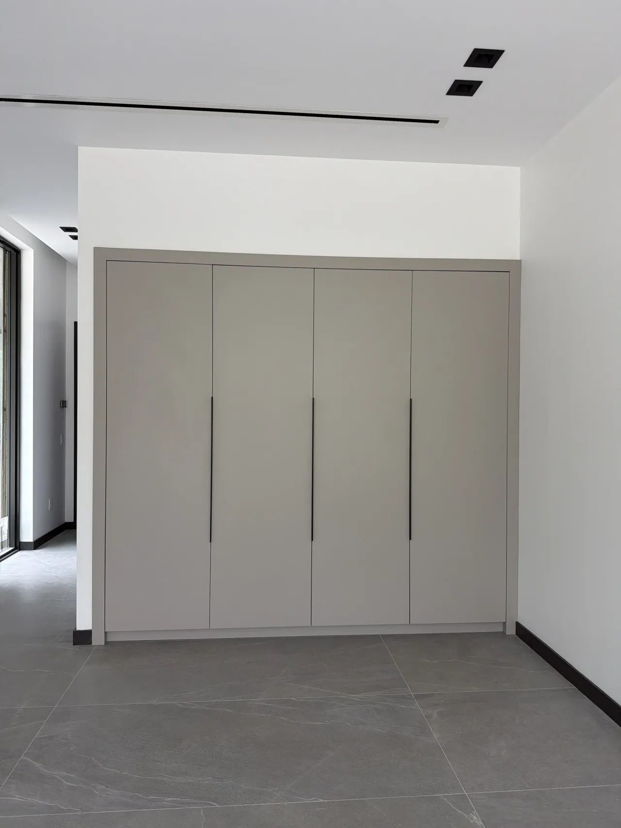 Closed greige four-door wardrobe with thin black bar handles set in a wall recess, in a bright room with a gray stone-look tile floor