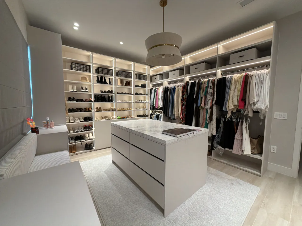 Closet combining shoe shelving, hanging clothes, a dresser, and a stone island with a bench in the foreground