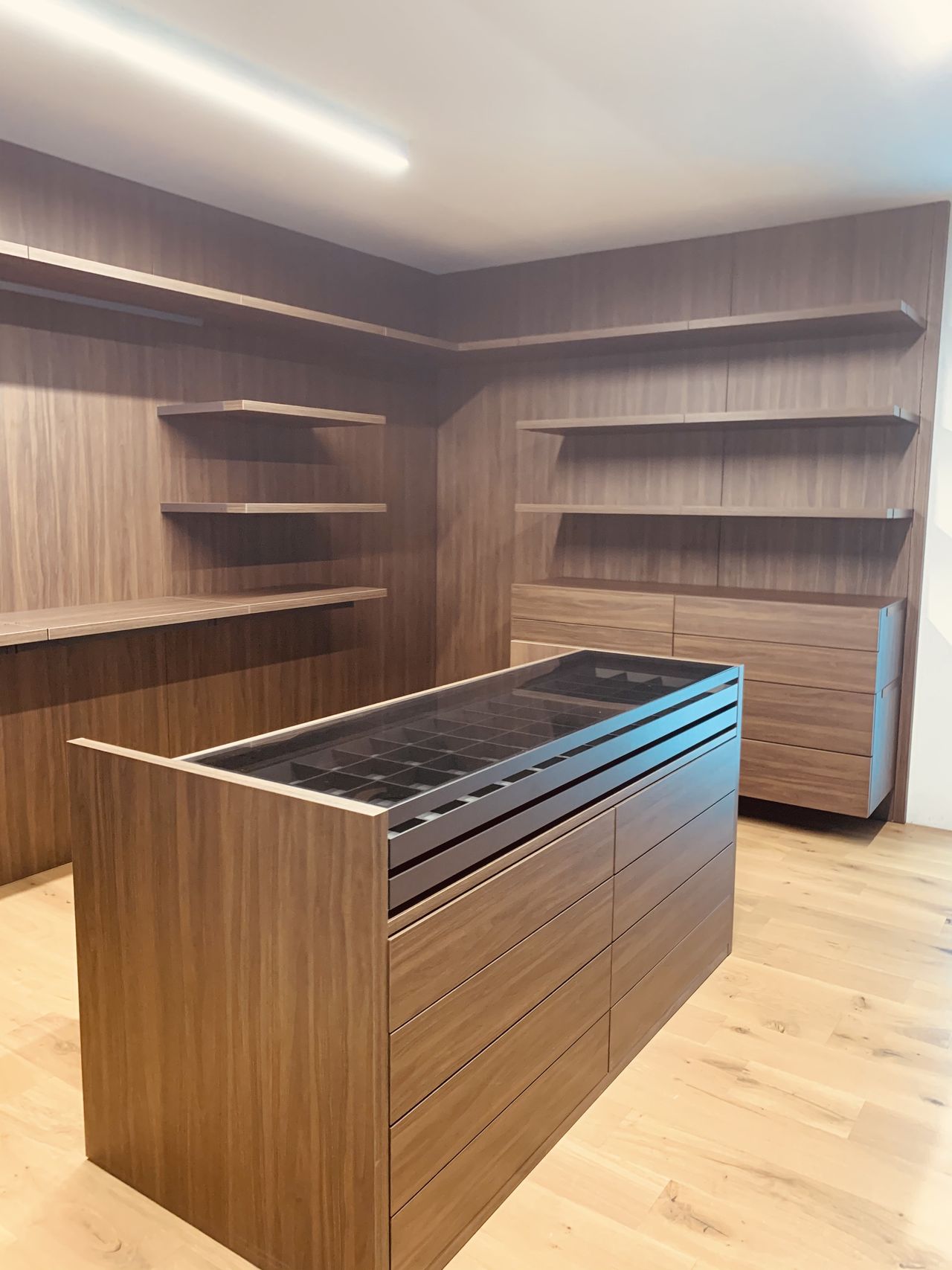 Closet corner in dark wood-grain with open shelving on two walls, drawer banks, and a center island with a divided glass-top tray over drawers