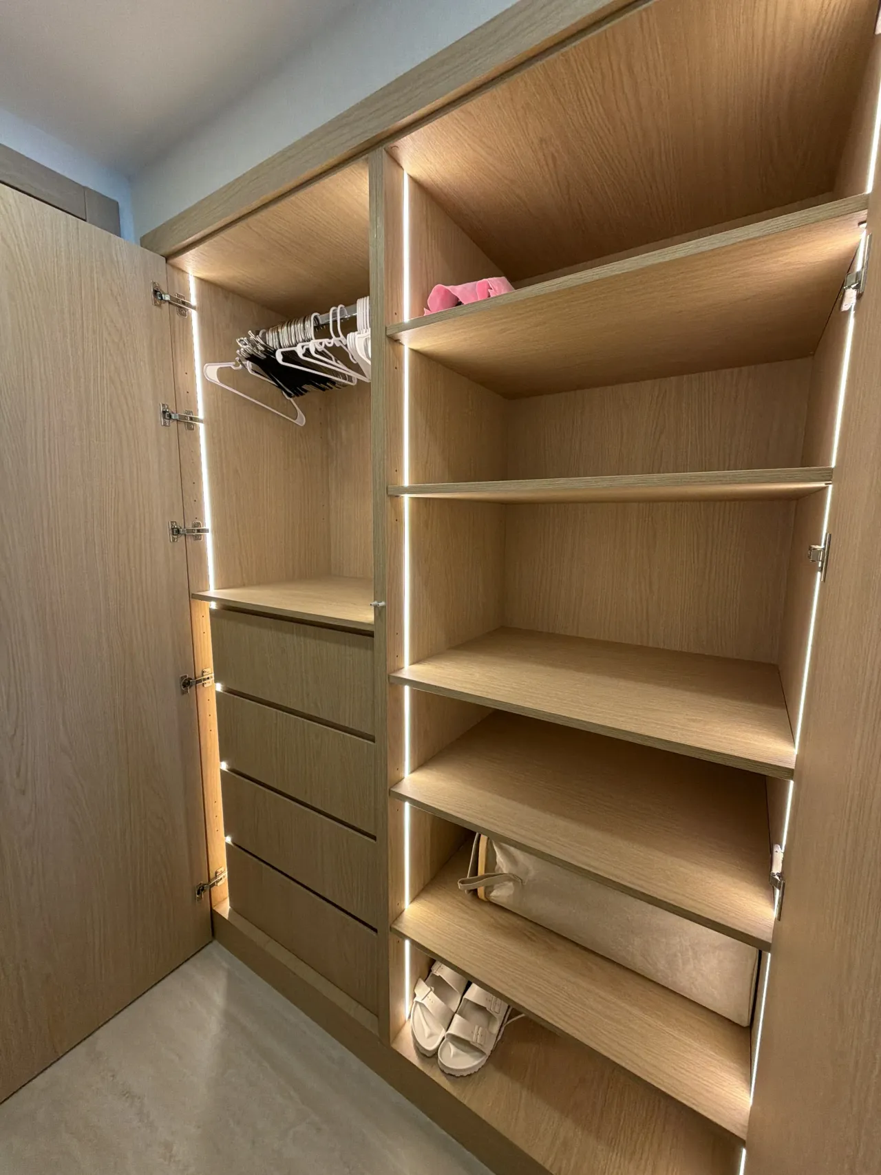 Closet corner in light wood-grain melamine with a rod of empty hangers and four drawers at left, LED-lit open shelves at right holding sandals