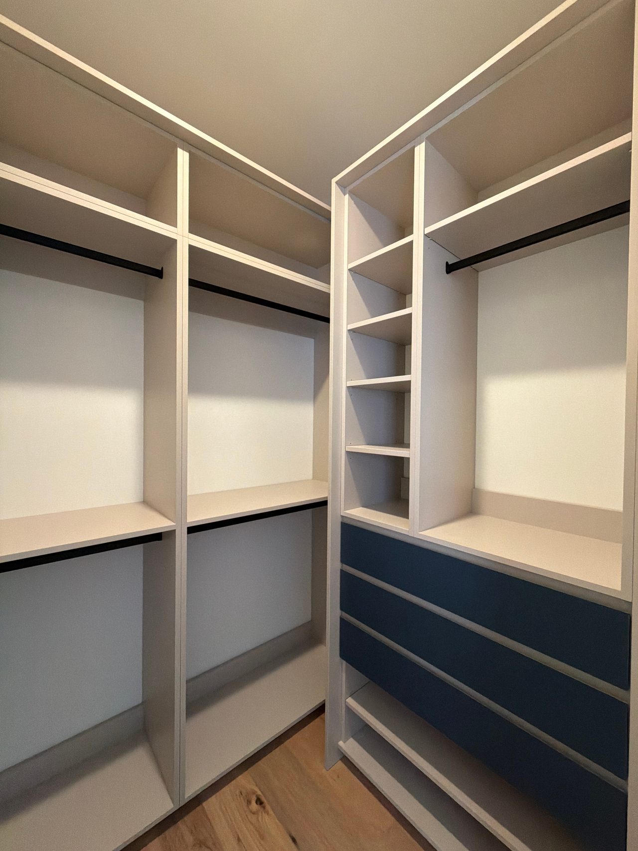 Empty corner walk-in closet with white open shelving, black hanging rods and a bank of navy blue drawers, over a light wood-grain floor