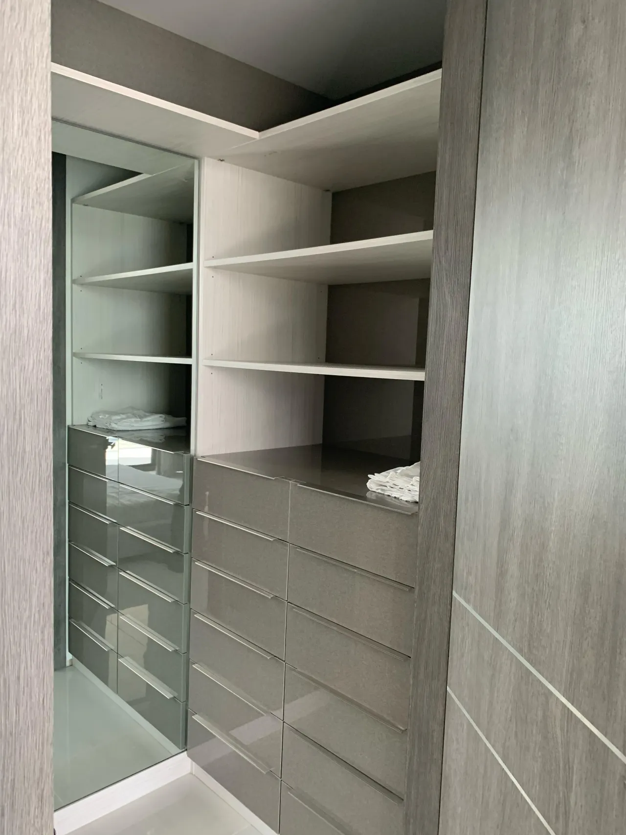 Closet corner with white open shelving above wide gray gloss drawer banks, a glass panel at left, and wood-grain gray side panels framing the run