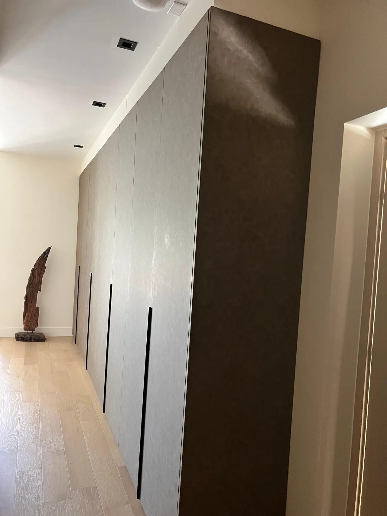 Corridor wardrobe run with gray textured door panels, thin dark recessed pulls and a dark brown end panel, light wood floor, sculpture at left