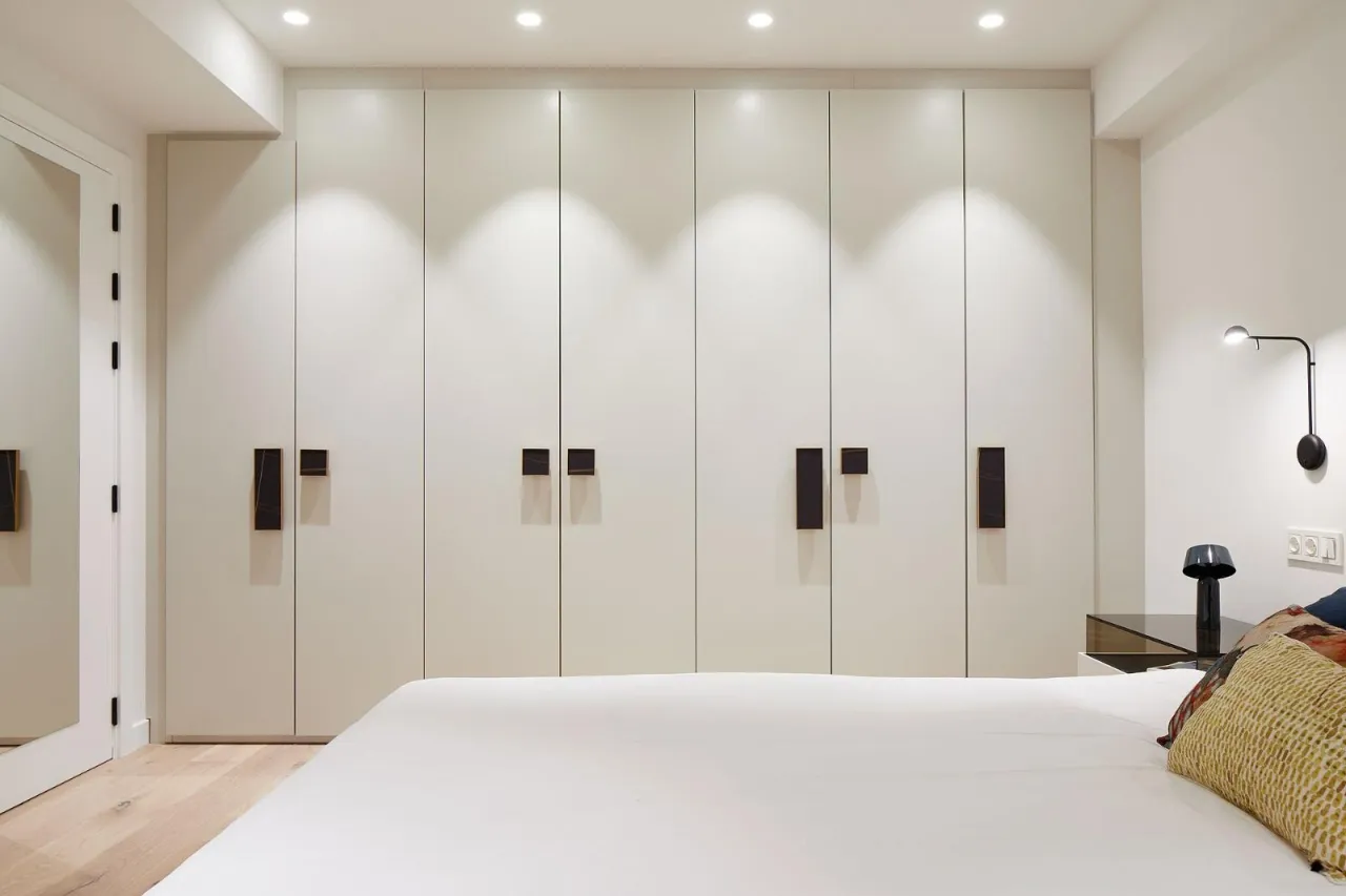 Cream built-in wardrobe wall with dark rectangular pull handles behind a bed, recessed downlights above, wall-mounted lamp and a mirror at left