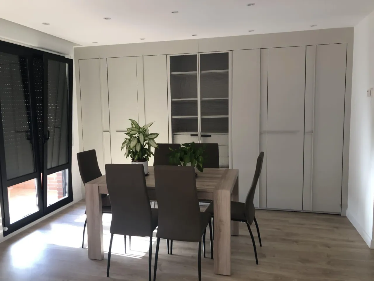 Cream floor-to-ceiling cabinet wall with a central pair of ribbed glass doors over shelves, flanked by handleless solid doors, dining table in front