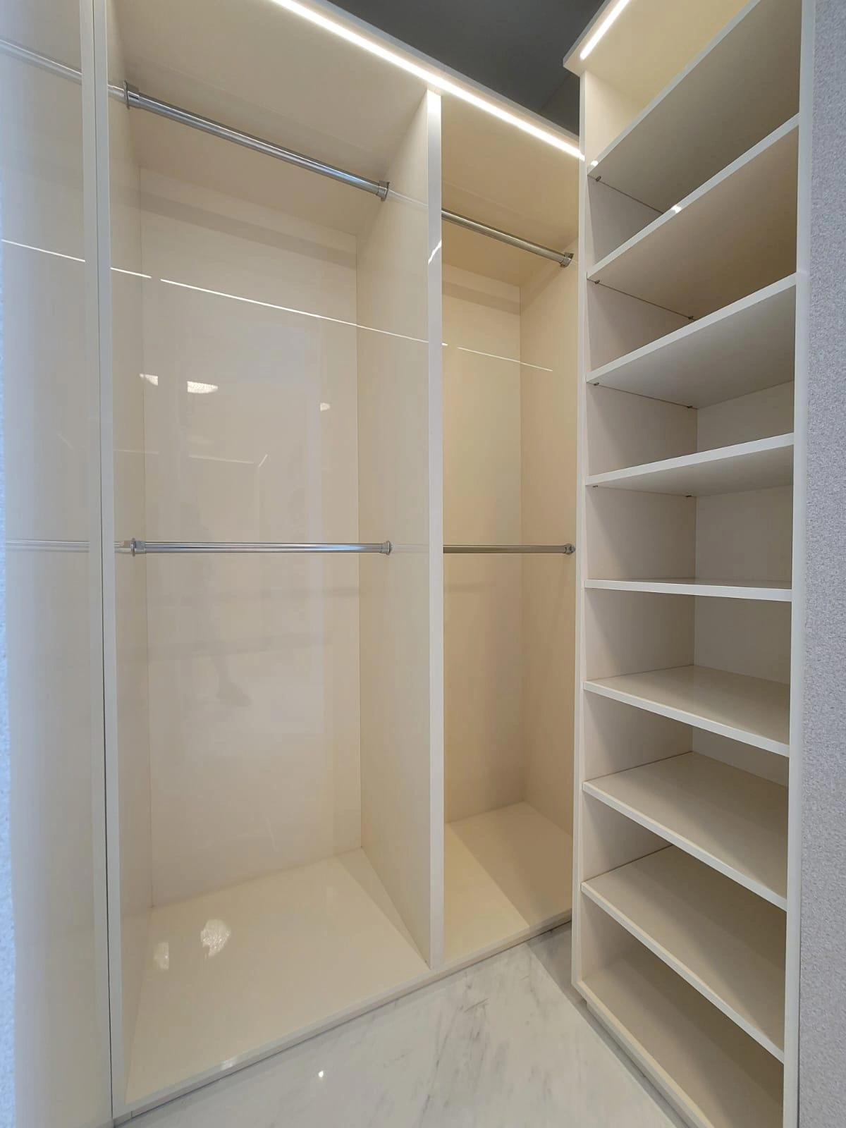 Cream high-gloss walk-in closet with two levels of hanging rods at left and a tall bank of angled open shelves at right, LED strip along the ceiling