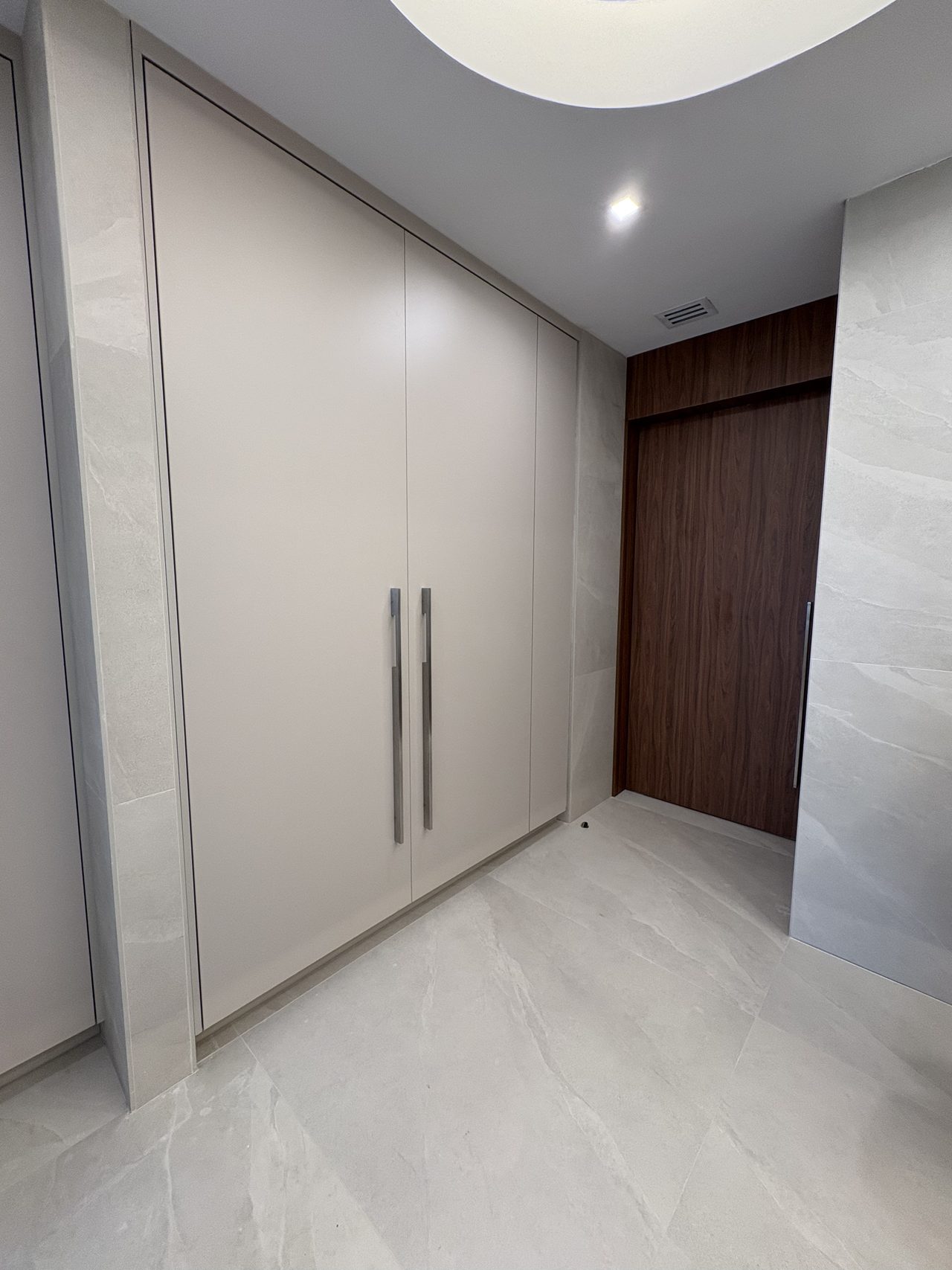 Cream/off-white flat-panel wardrobe with brushed metal bar handles in a hallway leading to a dark wood door, stone tile floor
