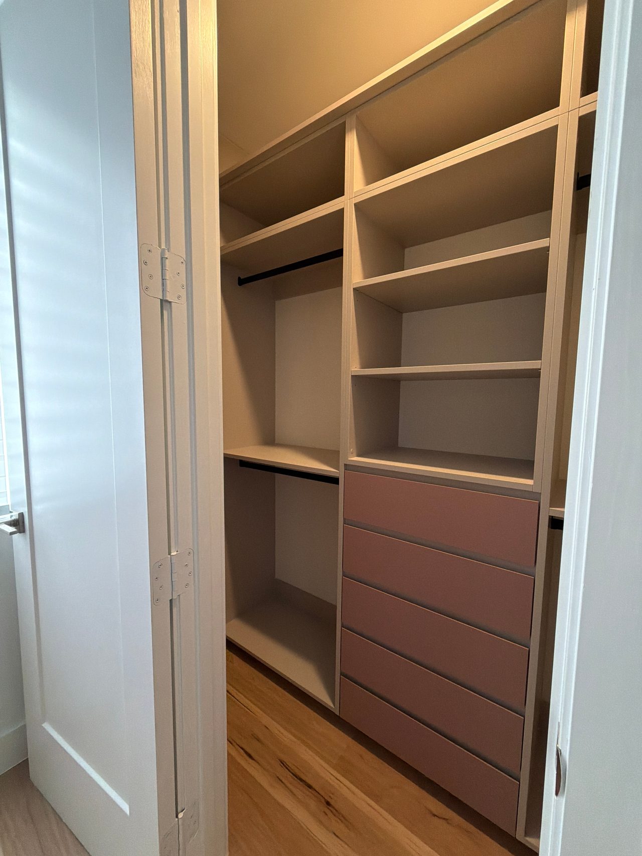 Closet interior with cream open shelving, black hanging rods and a five-drawer stack in mauve-pink beside a hinged door, over a wood-grain floor