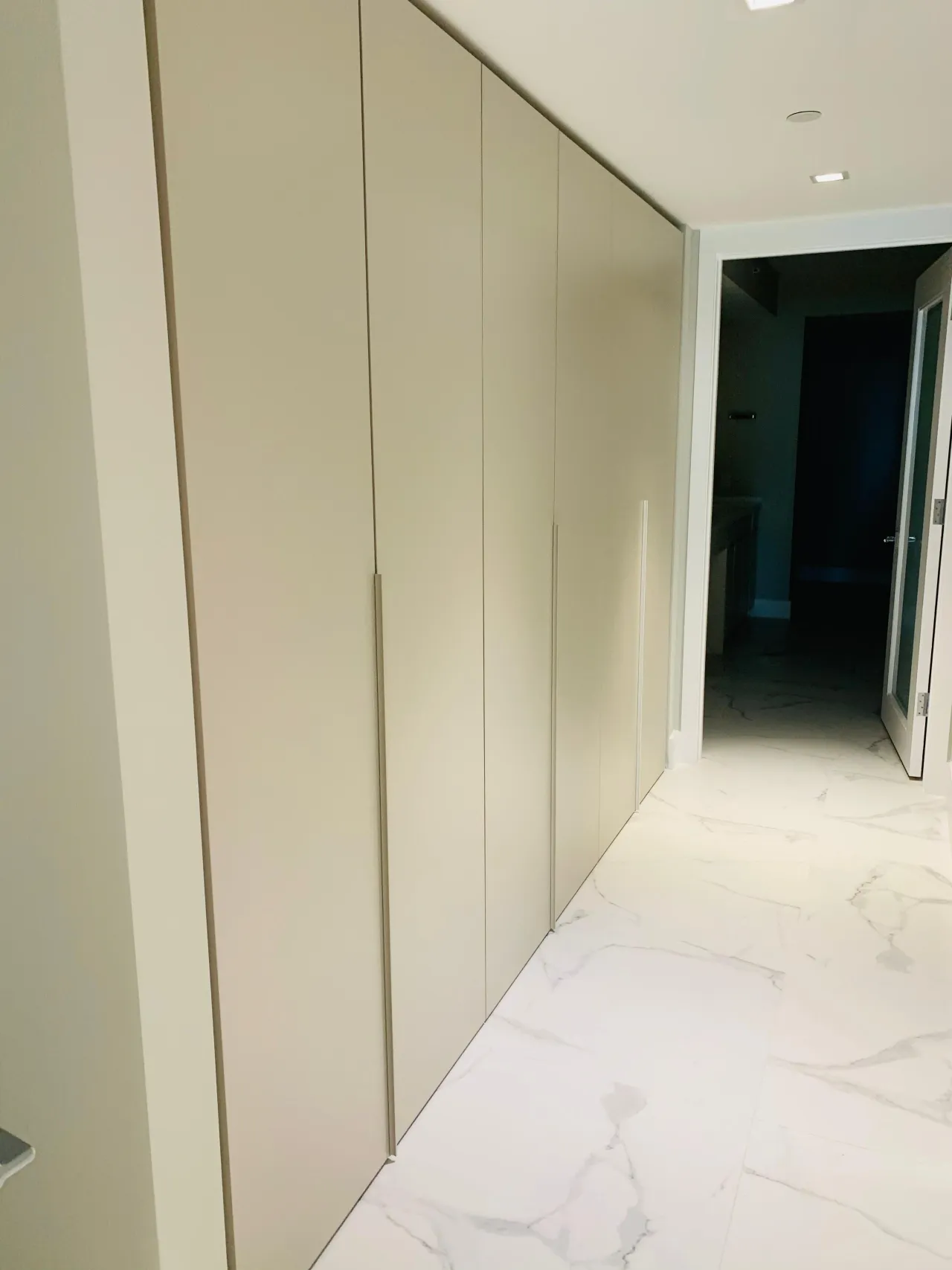 Cream/taupe floor-to-ceiling wardrobe with minimal vertical handles along a veined stone hallway leading to a dark doorway