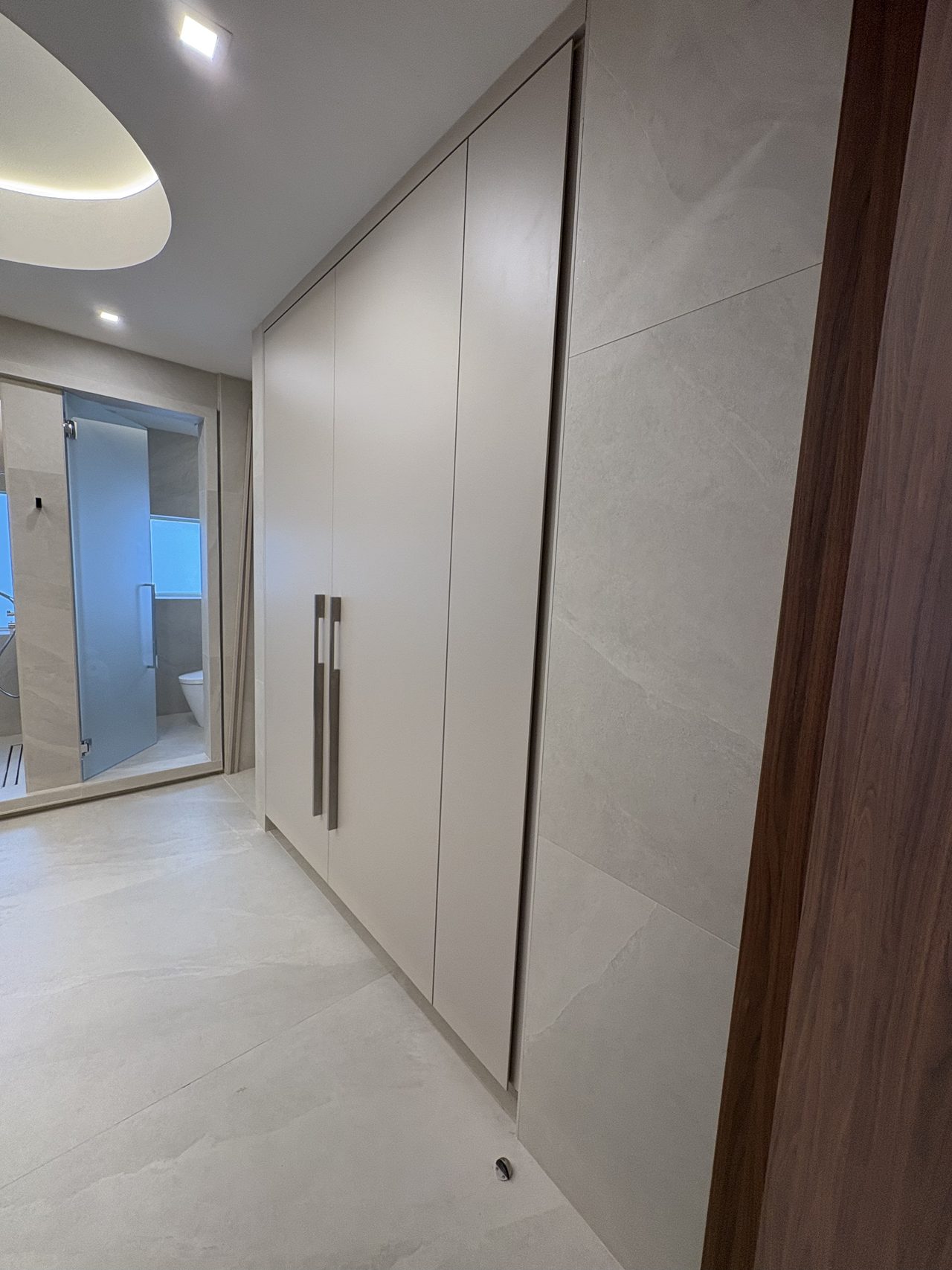 Cream wardrobe hallway leading toward a bathroom with a glass door and toilet visible