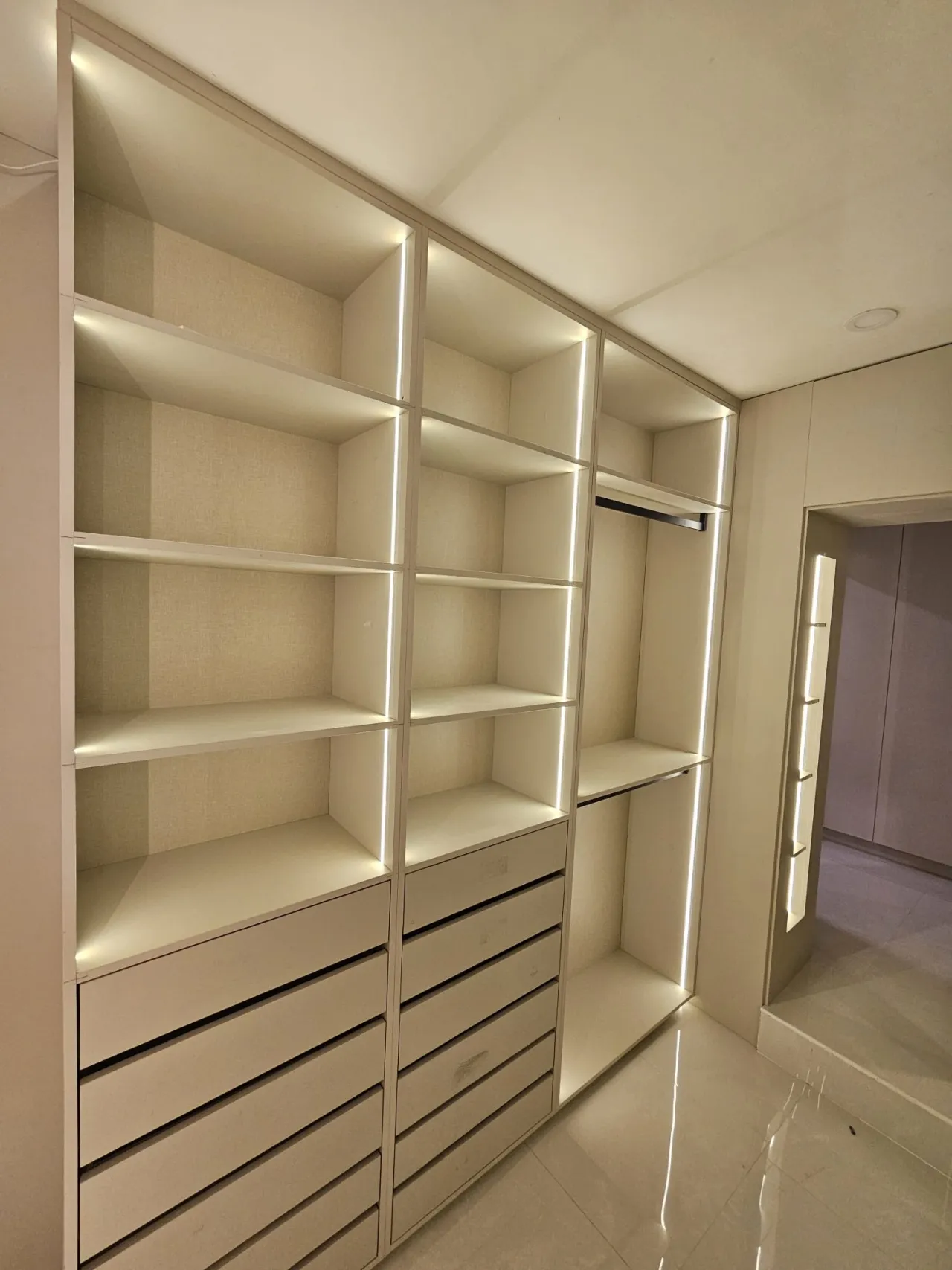 Cream/white walk-in closet with fabric-backed open shelving, drawer bank below, and LED edge lighting throughout