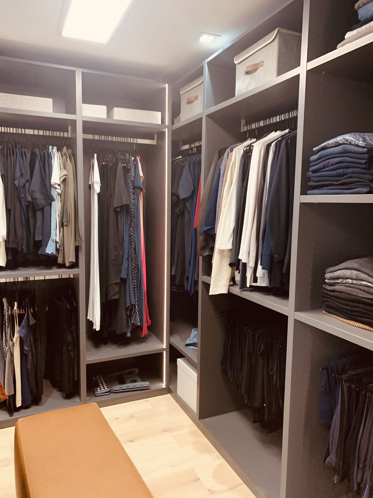 Dark charcoal walk-in closet with hanging clothes on both sides, lidded bins on top shelves, folded knitwear stacked at right, light wood floor