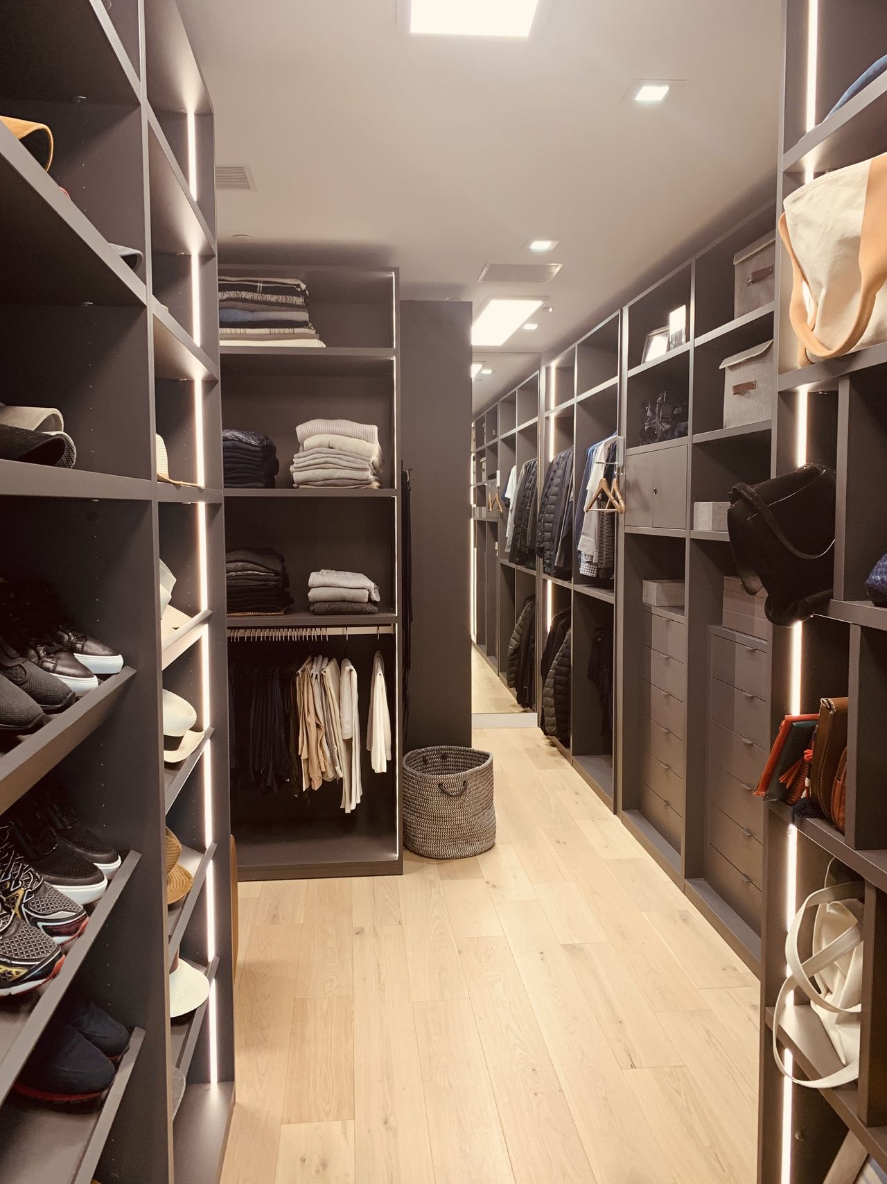 Dark gray-brown closet corridor with shoe shelving, folded clothes, hanging jackets, and a woven basket on a light wood floor
