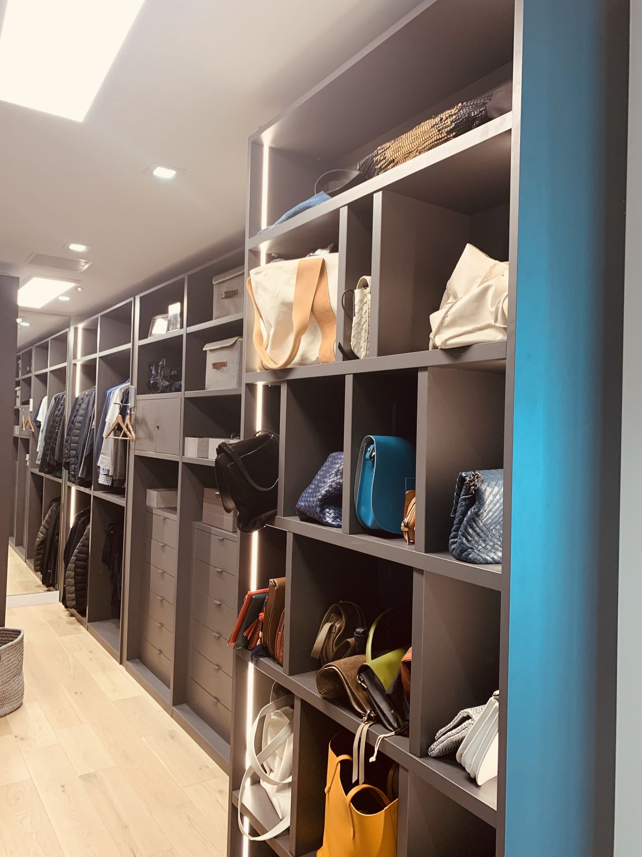 Long dark gray-brown open shelving closet displaying handbags and accessories with LED strip lighting and a blue accent wall, light wood floor