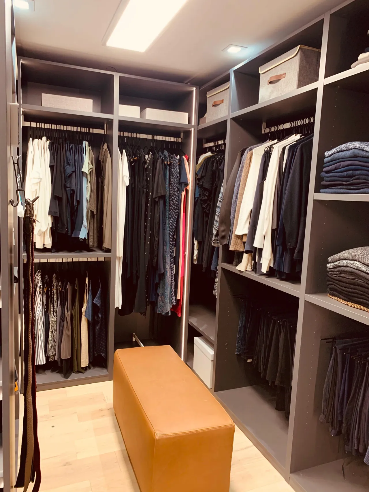Dark gray walk-in closet with double-hung clothing rods, storage boxes and folded stacks on upper shelves, and a tan upholstered bench in the center