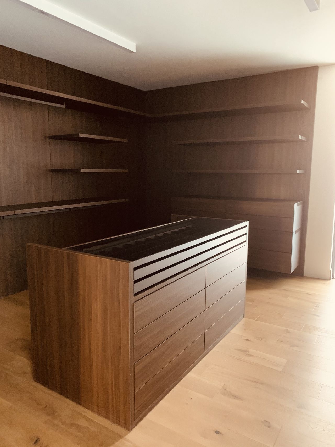 Dark wood closet corner with open shelving on two walls and a center island with a glass-top jewelry tray and drawers below, light wood floor