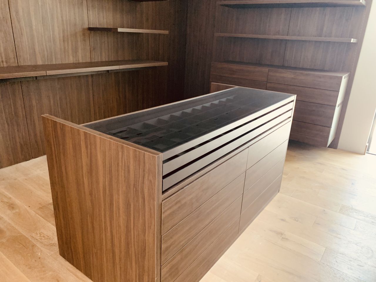 Dark wood closet island with a glass-top jewelry/watch tray insert above a bank of drawers, surrounded by matching dark wood shelving