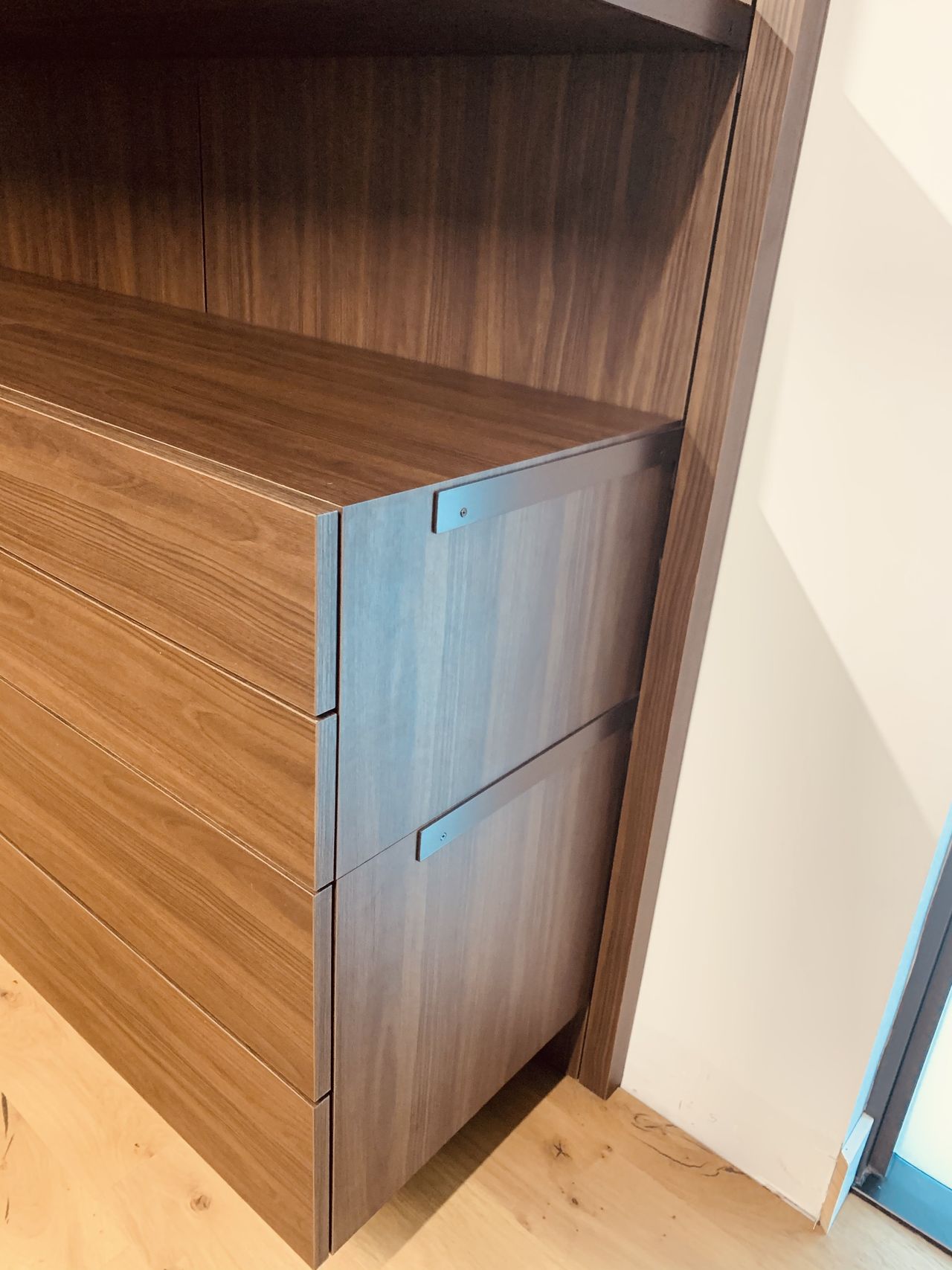 Two dark wood drawer fronts with small metal tab pulls set into a dark wood cabinet corner near a glass door frame