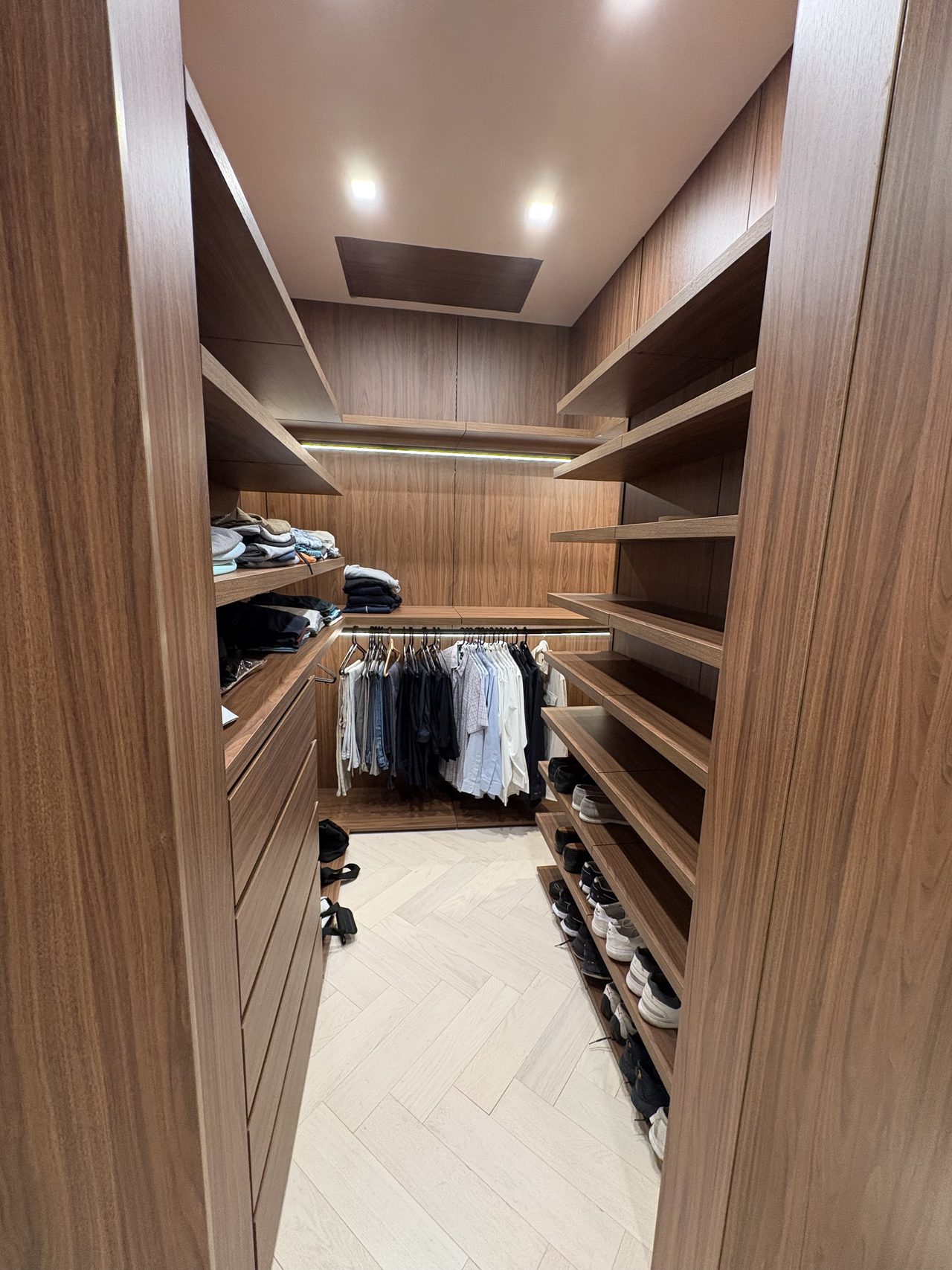 Dark wood-grain melamine closet aisle with drawers, hanging clothes, and shoe shelving on a herringbone floor