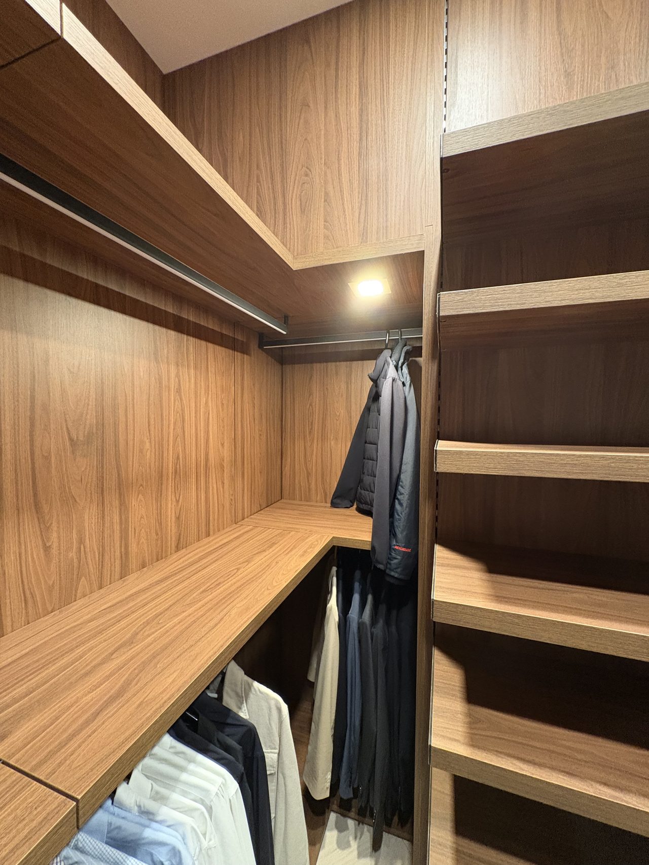 Dark wood-grain melamine closet corner with a hanging jacket, an L-shaped shelf, empty open shelving, and hanging shirts and trousers below