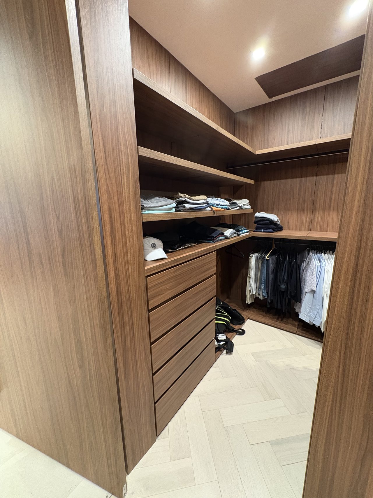 Dark wood-grain melamine closet with a drawer stack, angled shelves of folded clothes, and a rod of hanging shirts along the back wall