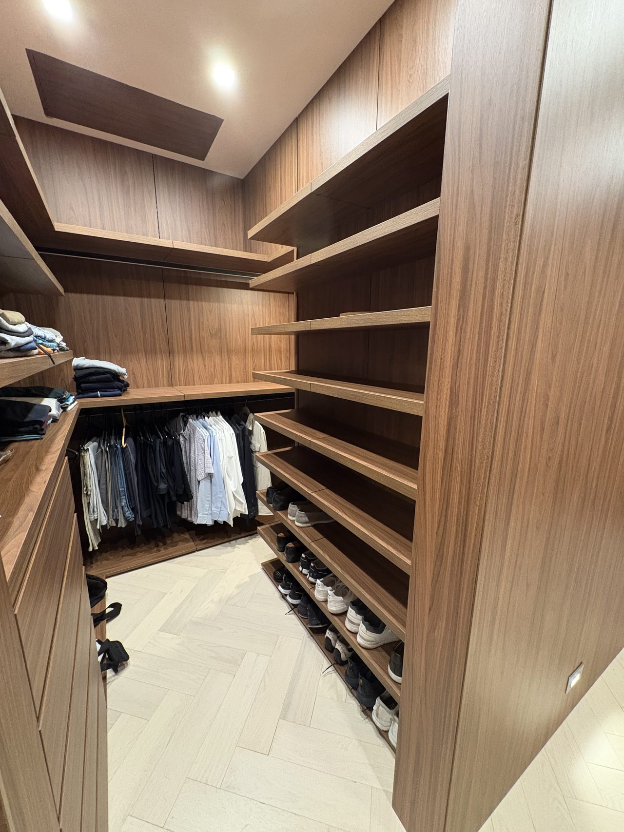 Dark wood-grain melamine closet with a tall column of open shoe shelves, a hanging rod of shirts, and a light herringbone-pattern floor