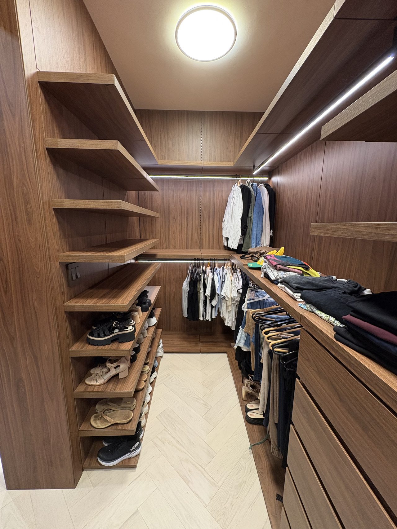Dark wood-grain melamine walk-in closet with diagonal display shelving, shoe shelves, hanging clothes, and a herringbone-pattern light floor