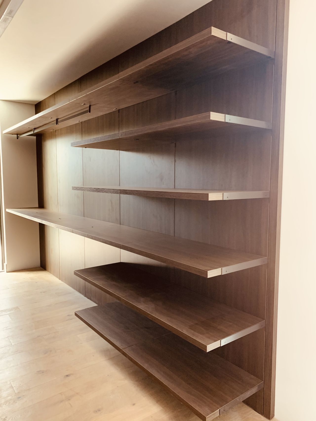 Dark wood-grain wall unit with horizontal floating shelves and a hanging rod above, empty, over a light wood plank floor