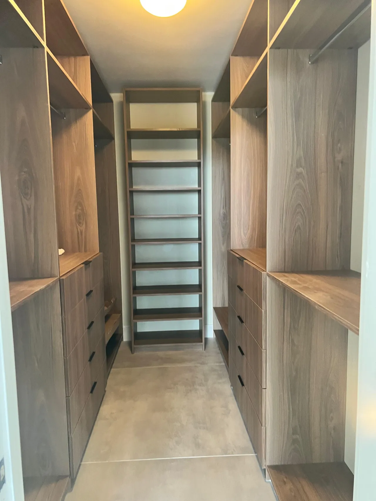Narrow dark wood walk-in closet with an end-wall shoe ladder shelf unit flanked by open shelving and drawer stacks, warm ceiling light