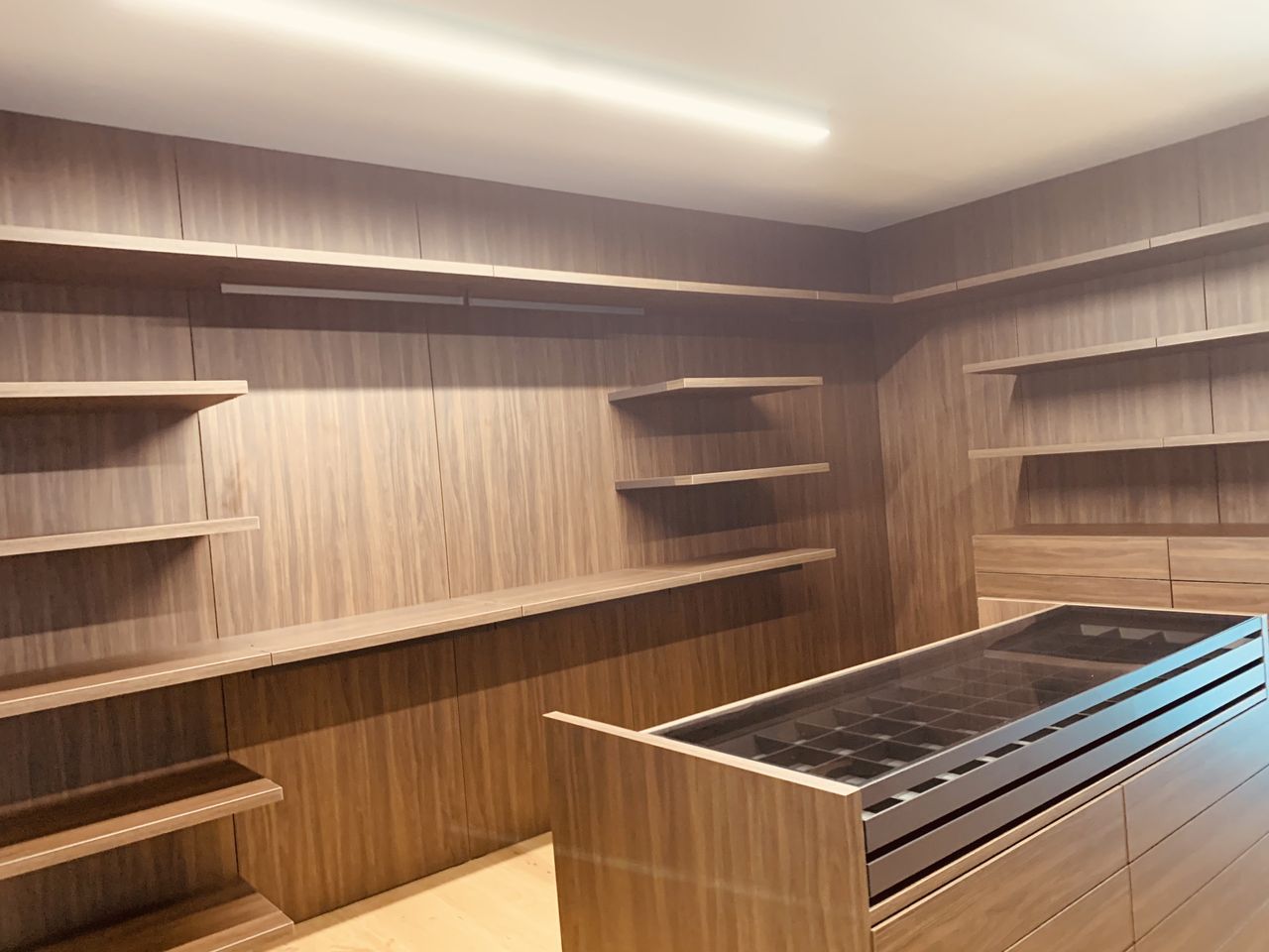 Dark wood wraparound open shelving with a horizontal hanging rod and a center island featuring a glass-top compartment tray and drawers, light wood floor