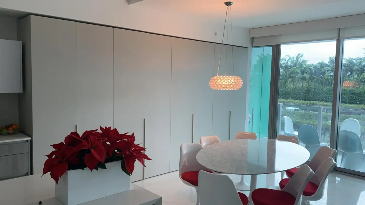 Dining area with a cream wardrobe wall behind a round stone table set, a modern pendant light, and greenery visible through glass sliding doors