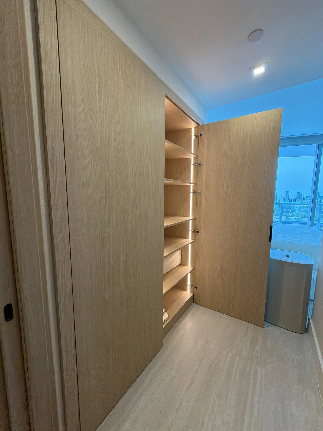 Closet with one door open showing LED-lit shelves in light wood-grain melamine, next to a floor-to-ceiling window with a city view