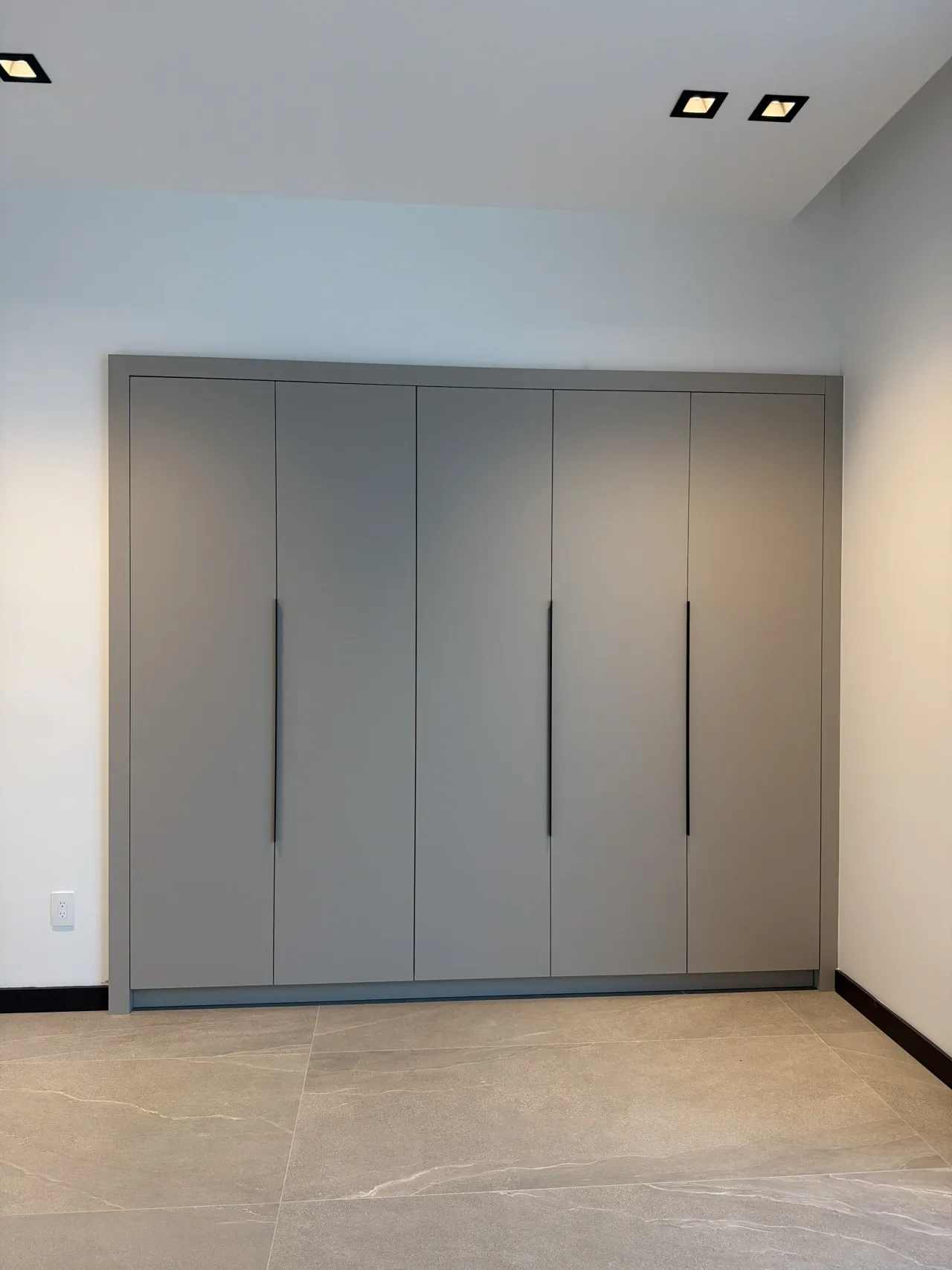 Five-door gray flat-panel wardrobe wall with slim black vertical pulls, running floor to ceiling on a beige stone-look tile floor