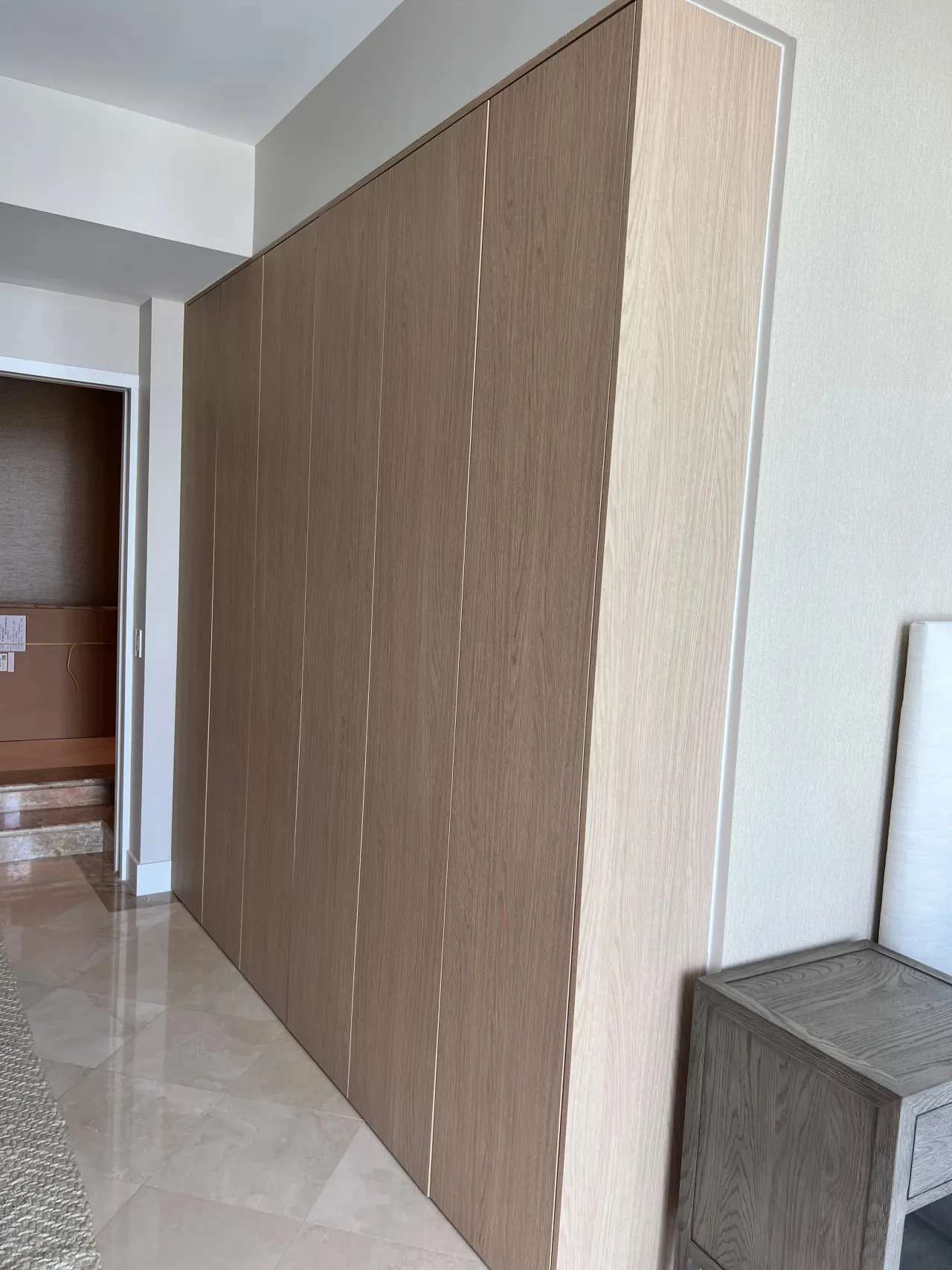 Floor-to-ceiling handleless wardrobe doors in light wood-grain along a hallway, slim recessed reveals between panels, glossy stone-look tile floor