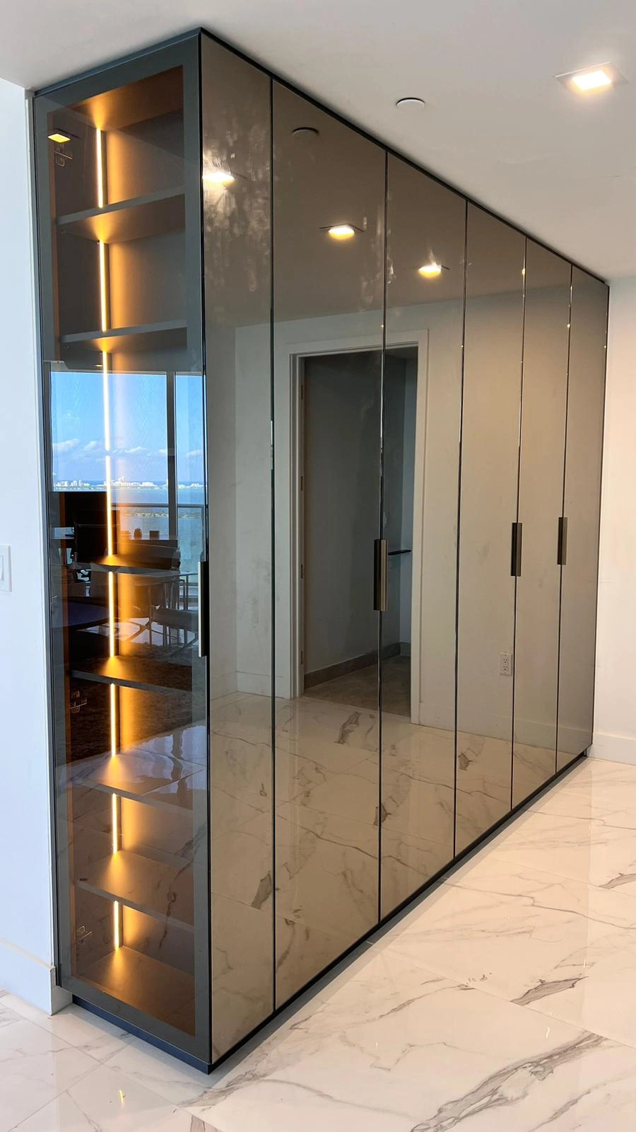 Floor-to-ceiling wardrobe with dark tinted glass fronts and slim metallic vertical pulls, LED-lit open shelving at one end, veined stone tile floor
