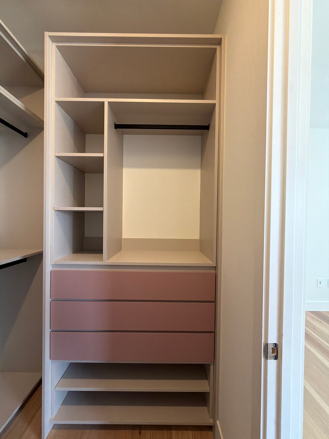 Freestanding closet unit with open upper shelves, a black hanging rod, three pink drawer fronts and two open shelves at the base