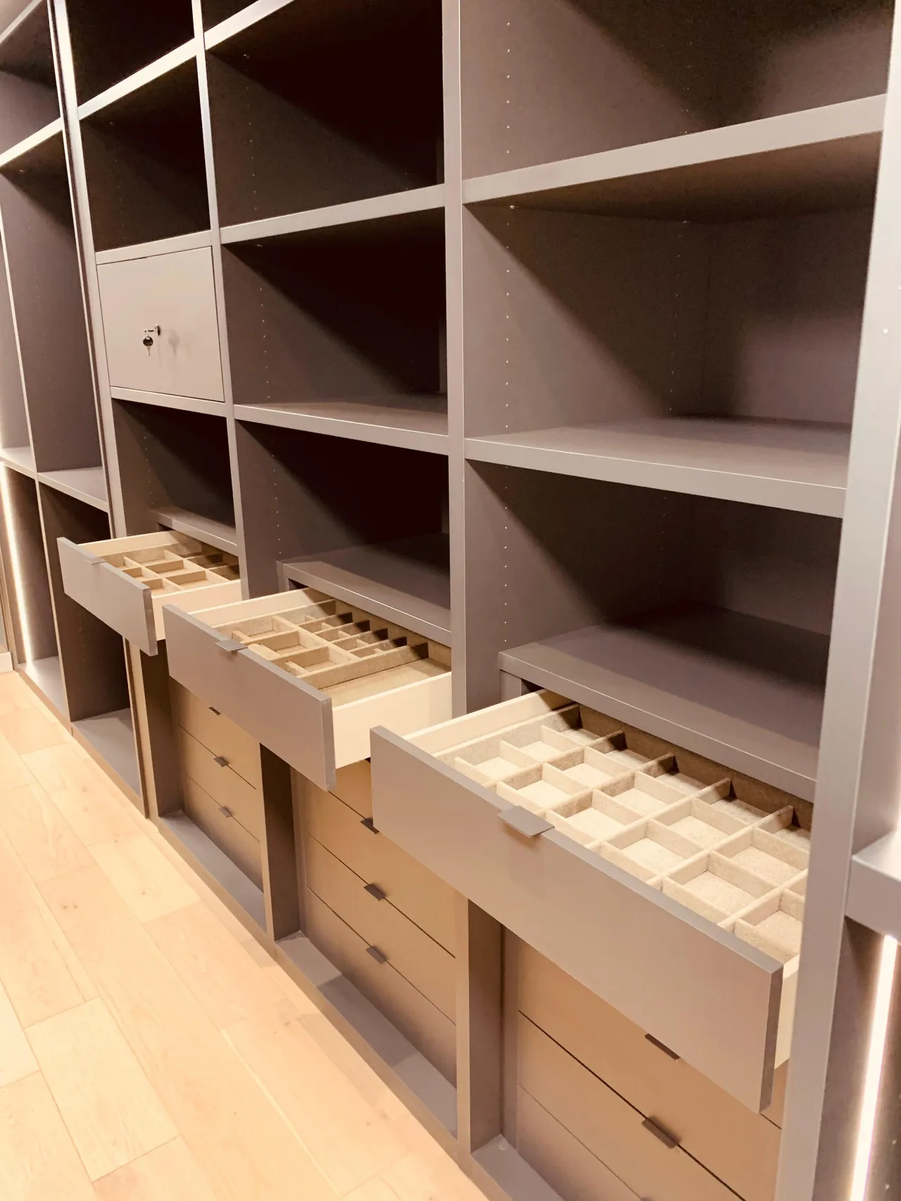 Gray/taupe closet interior with open drawers showing felt-lined compartments for jewelry, and a small built-in safe on an upper shelf