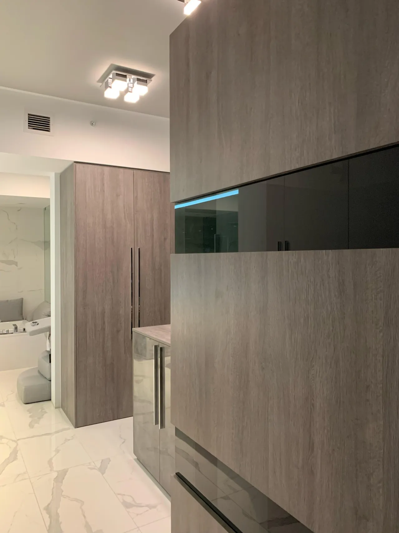Gray wood-grain wardrobe with a black glass upper display cabinet section, next to a mirrored dresser piece, on a white stone floor