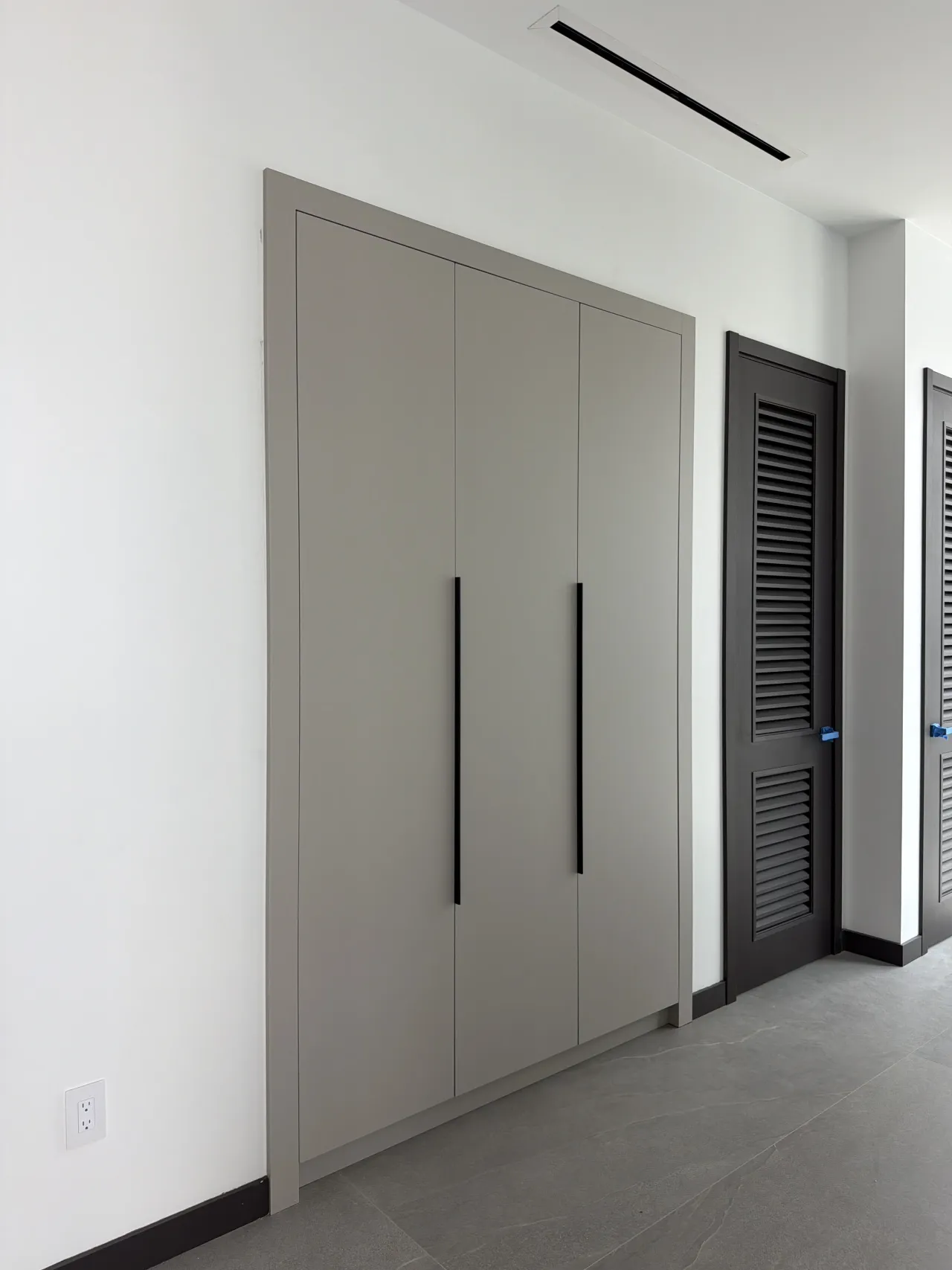 Greige three-door wardrobe with black bar handles beside a dark louvered closet door in a hallway with a gray tile floor