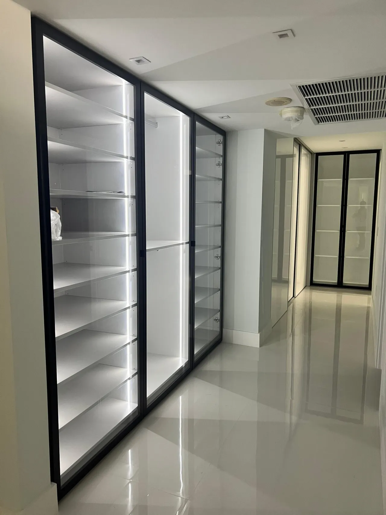 Long hallway of black-frame glass display cabinets with LED-lit shelving on both sides, glossy stone floor extending toward more glass doors