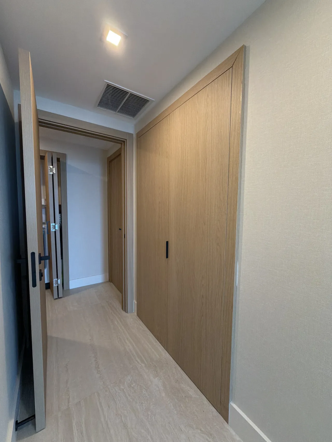 Hallway with floor-to-ceiling light wood-grain melamine closet doors with slim black pulls, an open door at left and light stone-look tile floor