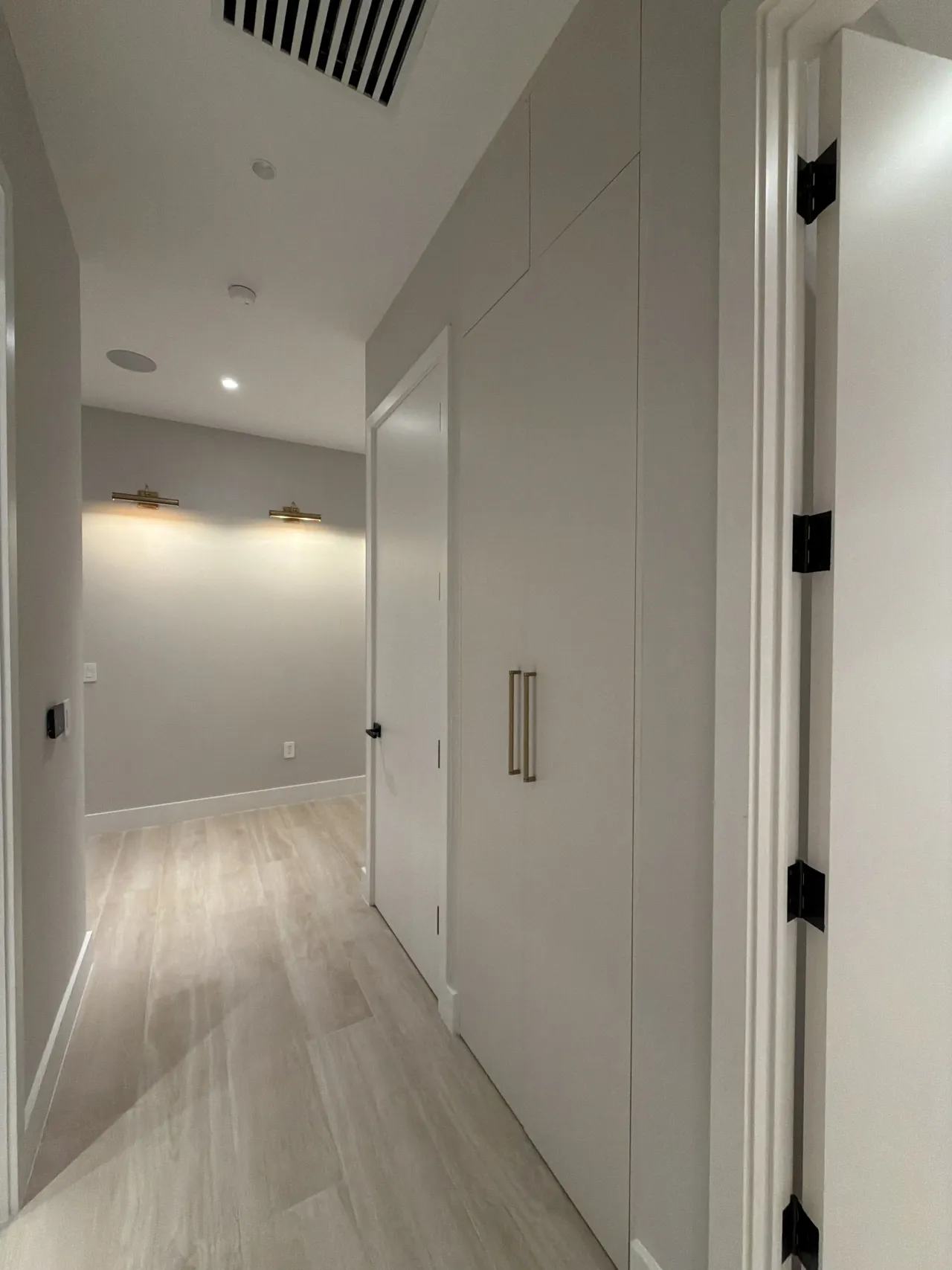 Hallway with a floor-to-ceiling off-white cabinet run with long gold-tone bar pulls, an open passage door, and two picture lights beyond