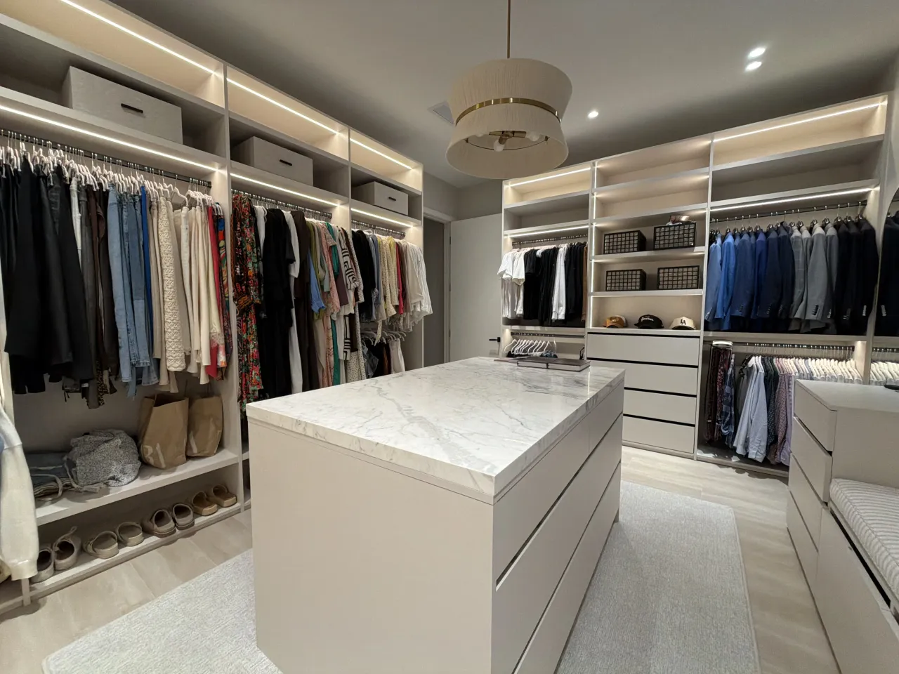 Closet with hanging clothes on both sides and a stone-top island in the center under a drum pendant light