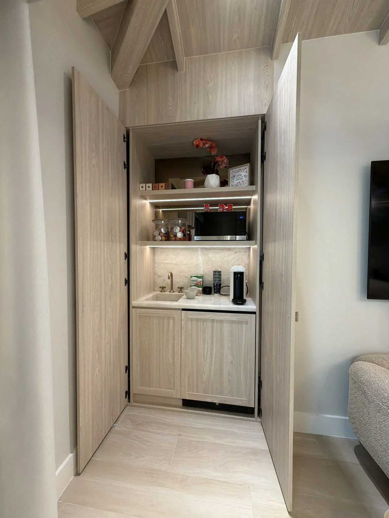 Kitchenette niche behind tall wood-grain melamine doors, with a sink, microwave, lit shelves, and a light stone backsplash and counter