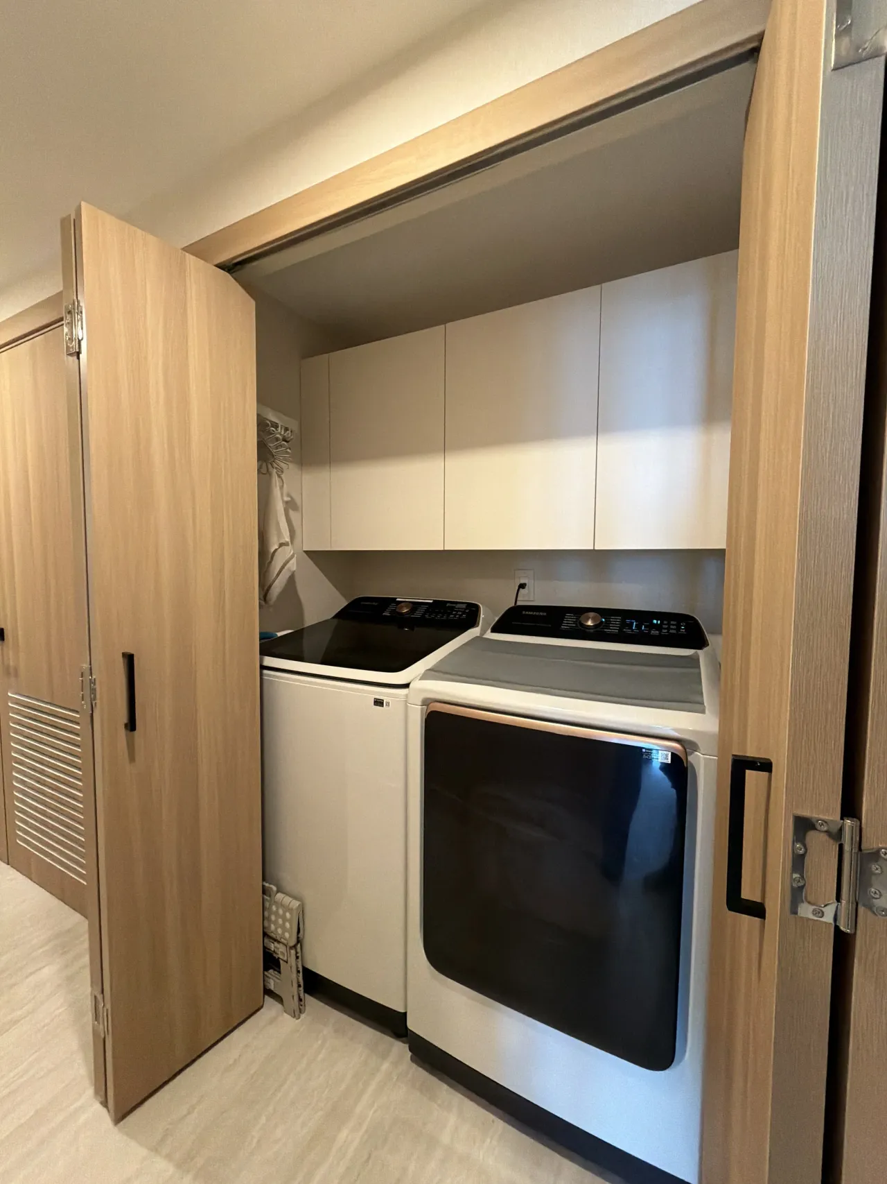 Laundry closet with a side-by-side white washer and dryer behind light wood-grain melamine folding doors, light upper cabinets above