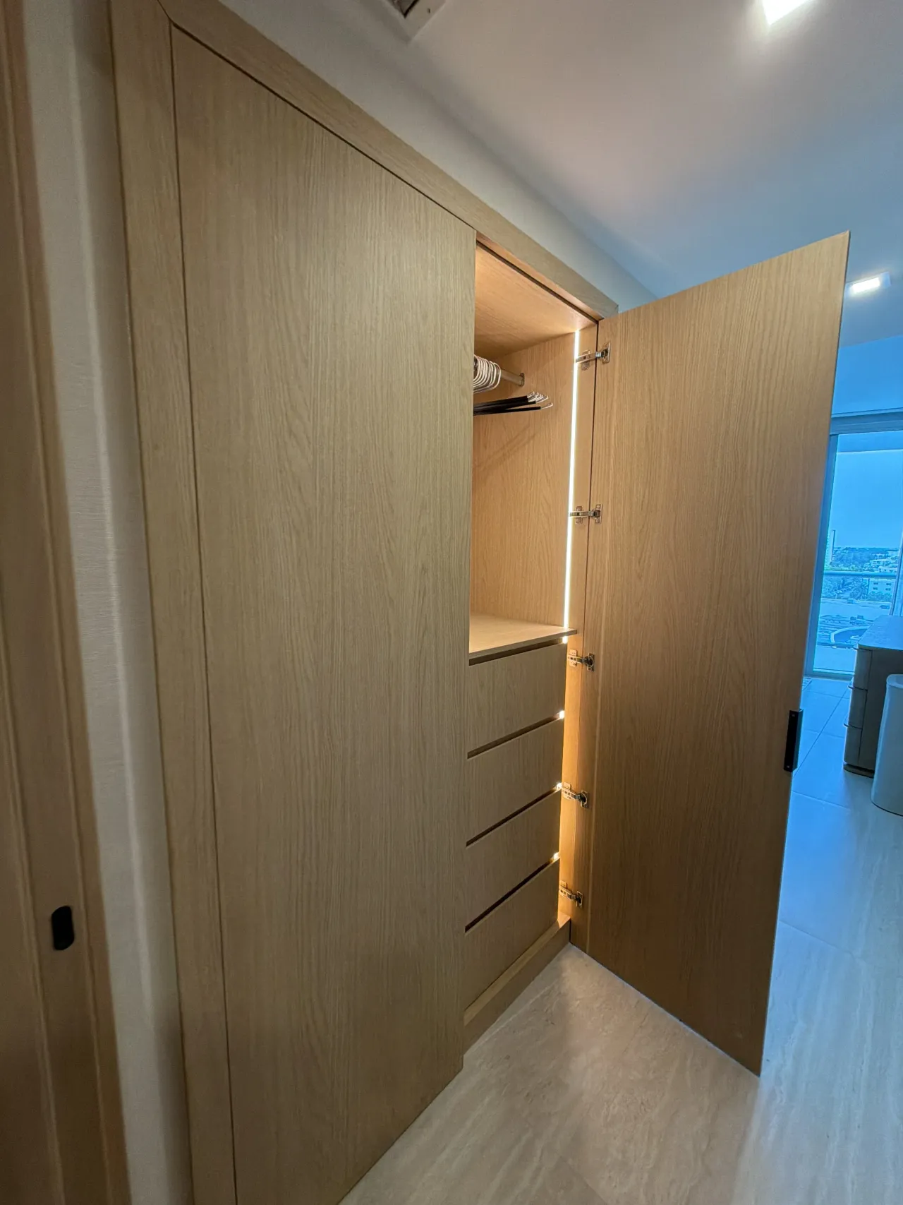 Light melamine closet with doors open revealing a lit interior shelf with a hanging rod and a drawer stack, city view window visible through the open door