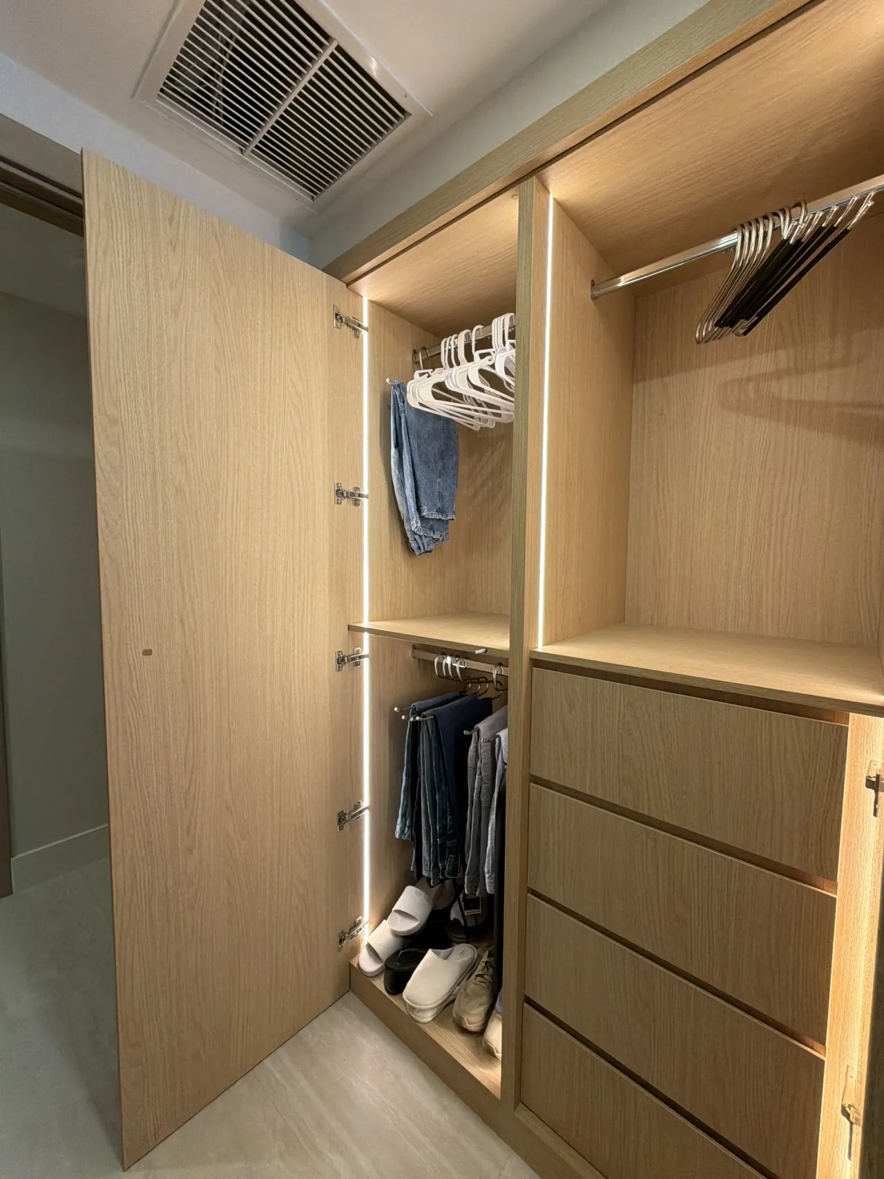 Light wood-grain melamine closet with one door open revealing hanging jeans, a shoe shelf, and a drawer stack with LED edge lighting