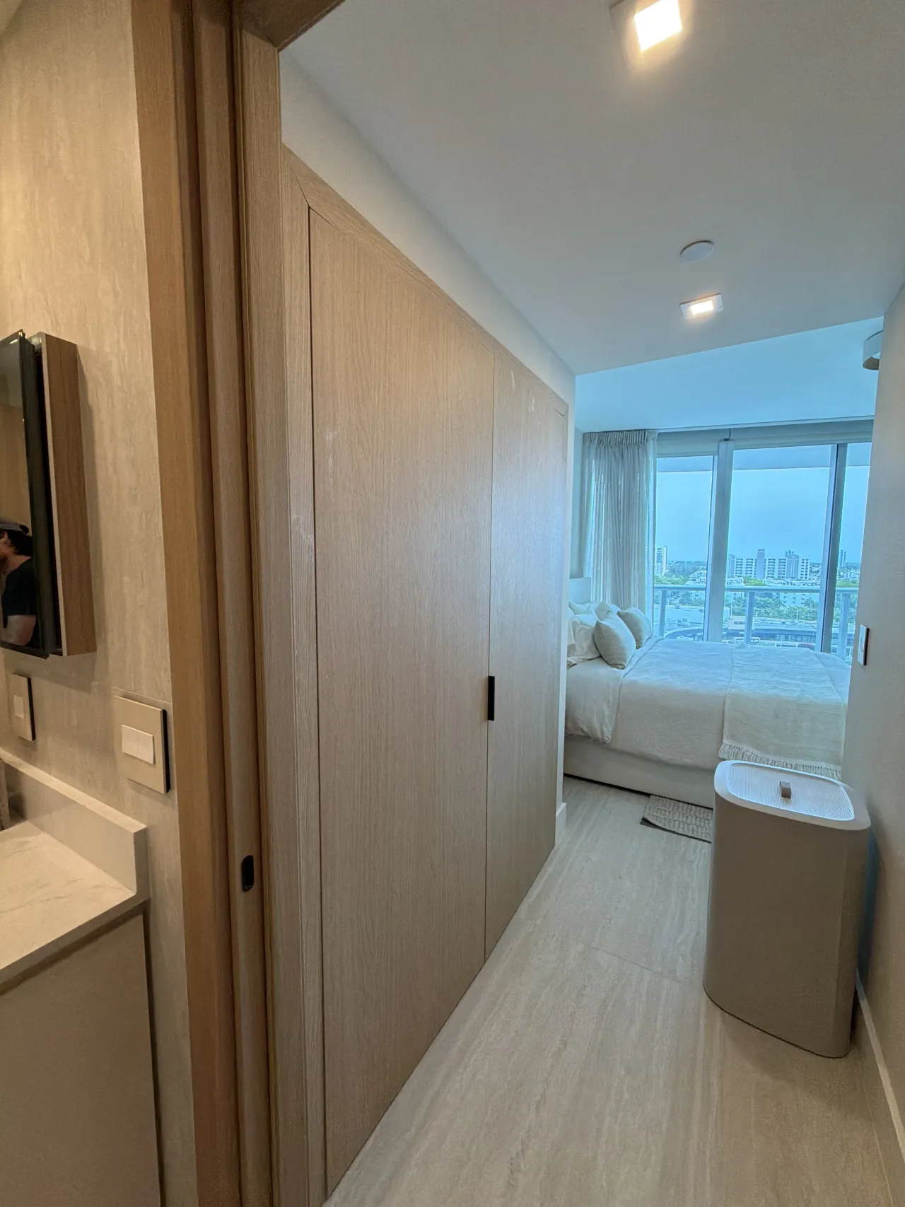 Light wood-grain melamine closet doorway opening onto a bedroom with floor-to-ceiling windows showing a city skyline view, light stone floor