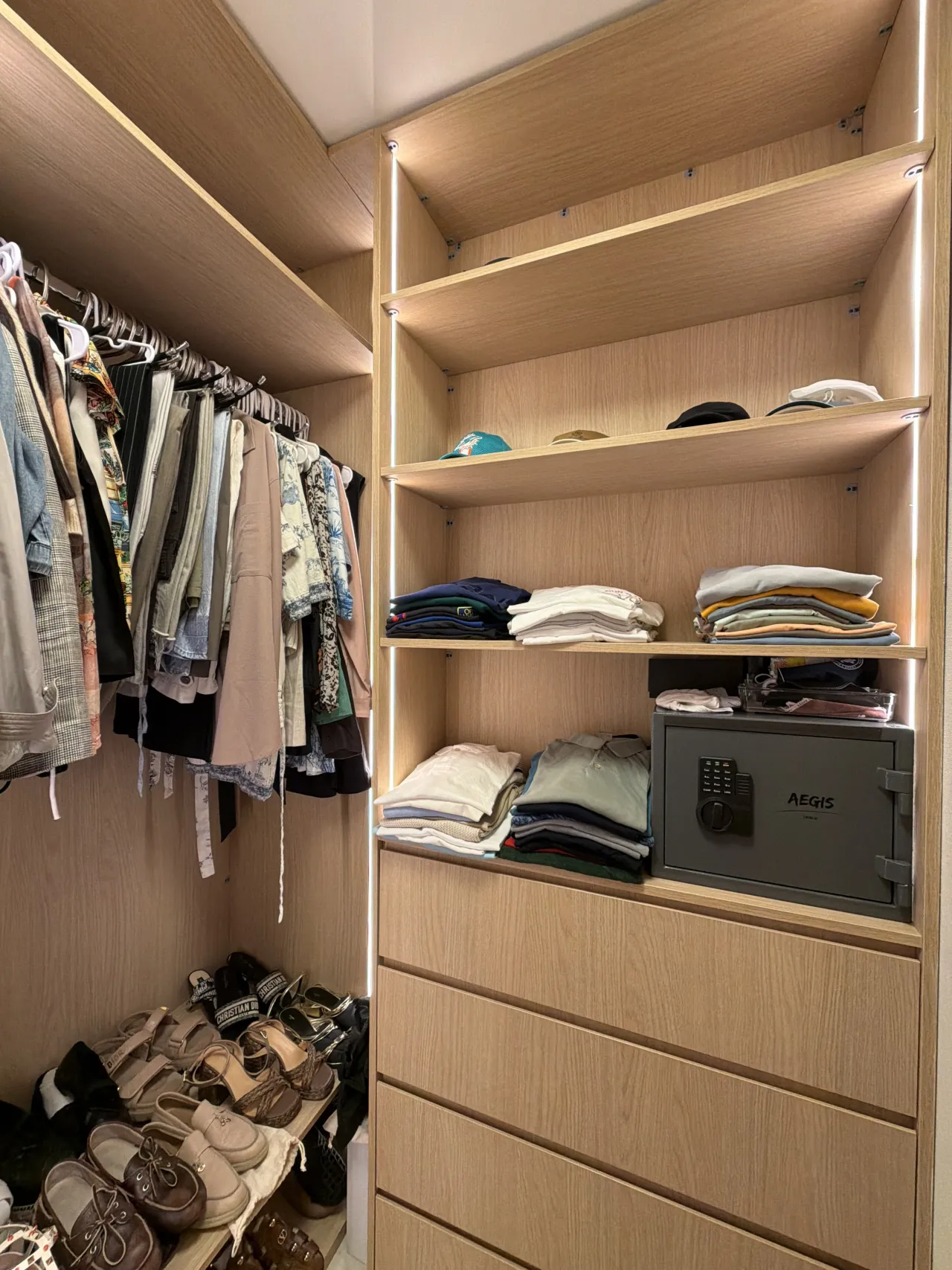 Light wood-grain melamine closet shelving with folded clothes, caps, and a gray floor safe set into a lower shelf, LED edge lighting