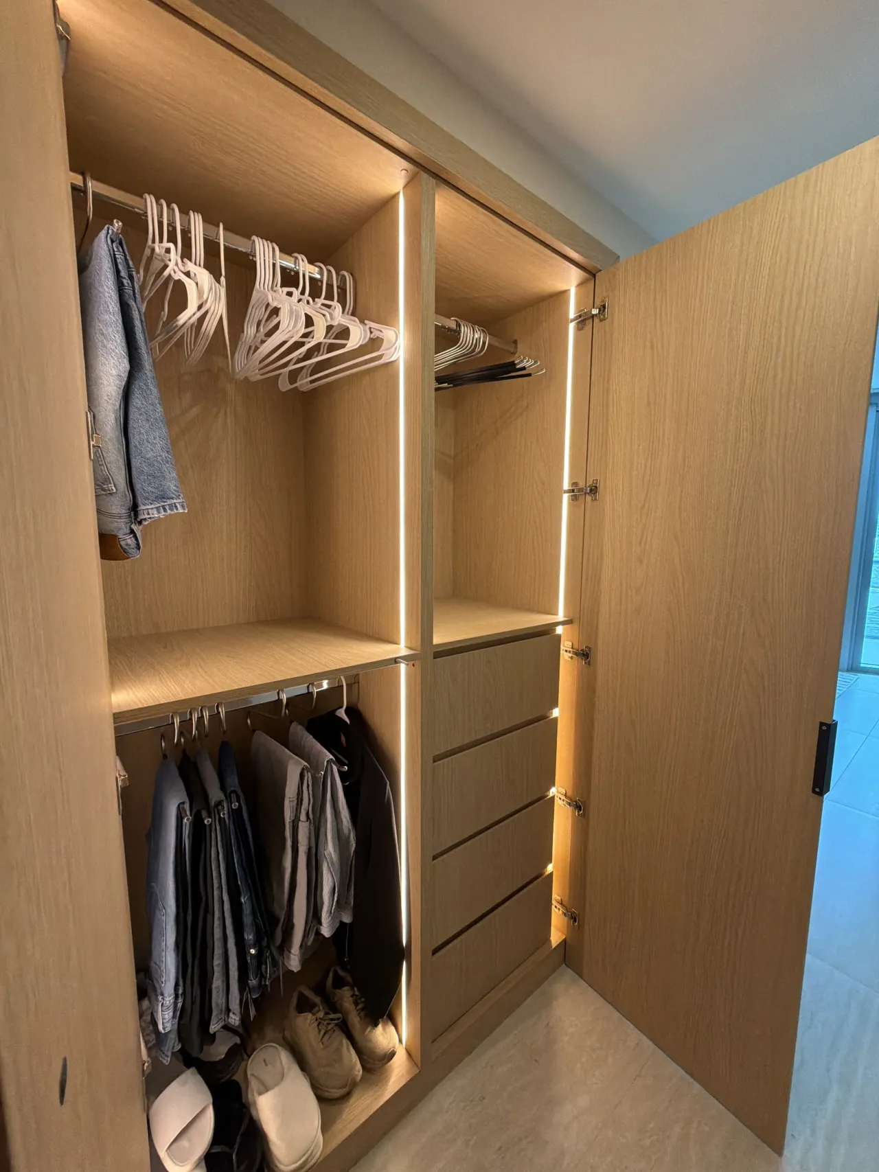 Closet interior in light wood-grain melamine with two hanging rods of jackets and shirts, LED strip lighting, handleless drawer stack and shoes below